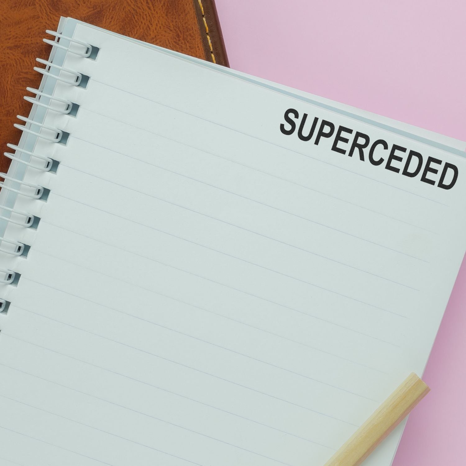 Spiral notebook page with the word superceded, a pencil, and a pink desk background.