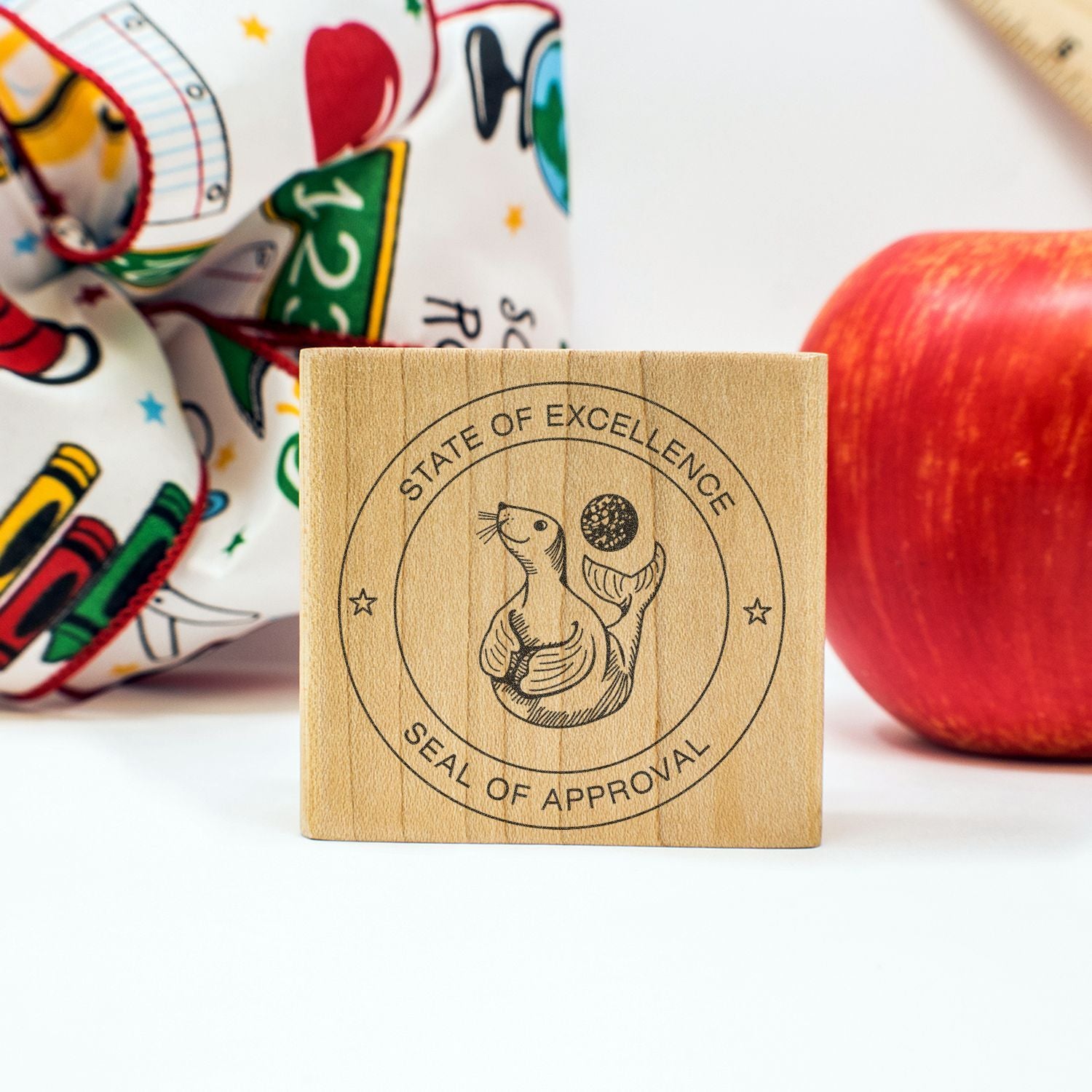 Wooden seal of approval stamp with State of Excellence design, apple, and colorful school supplies background