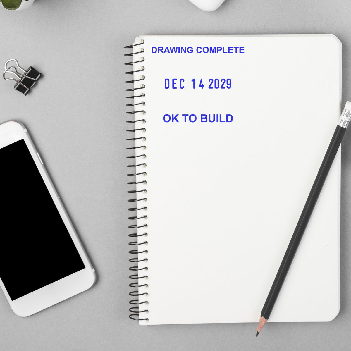 Notebook with Drawing Complete, Dec 14 2029, and OK to Build text beside a pencil, phone, and binder clip on a desk.
