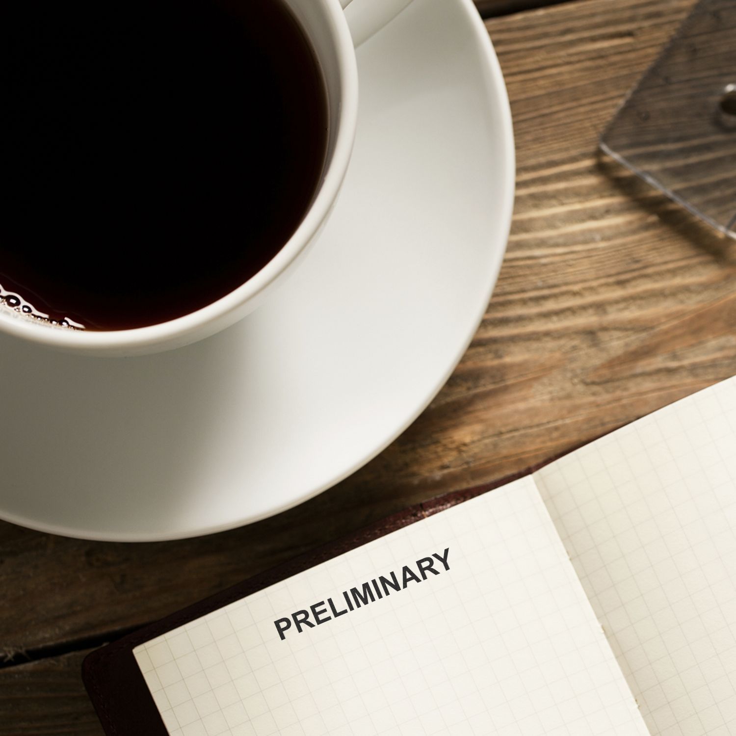 Black coffee cup beside an open notebook labeled Preliminary on a wooden desk, minimal office workspace scene.