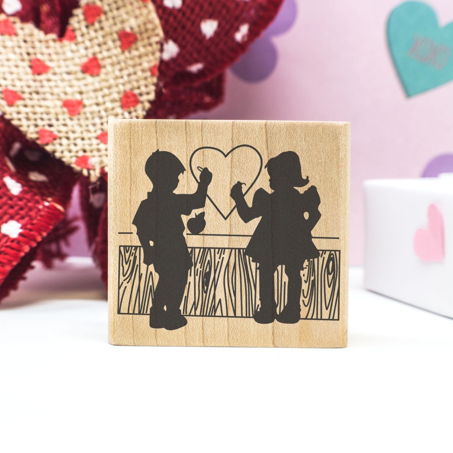 Wooden Valentines Day stamp with boy and girl silhouettes drawing a heart, handmade craft gift on a pastel background.