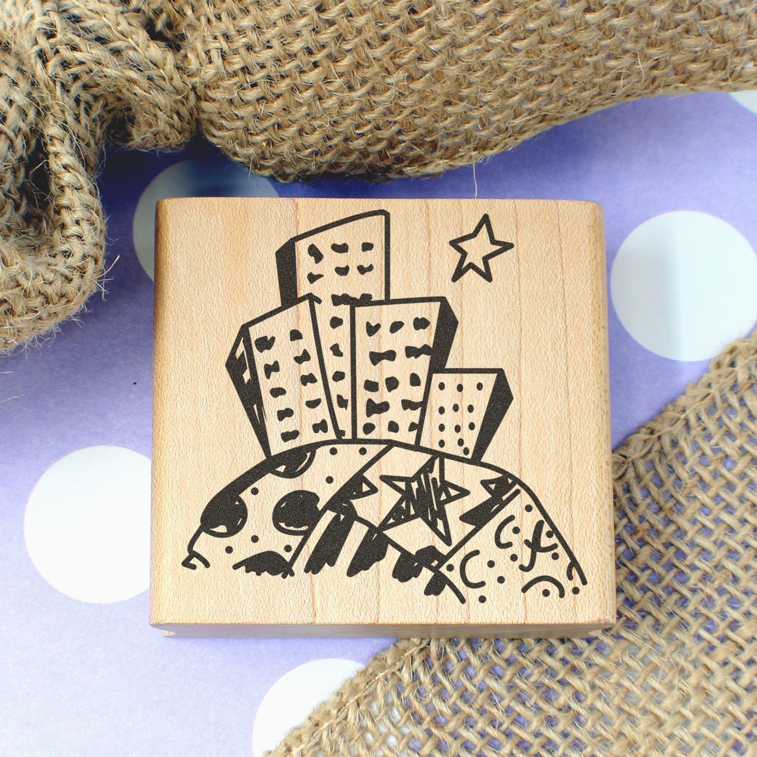 Wooden rubber stamp with whimsical city skyline illustration on purple polka dot background, rustic craft supply close-up