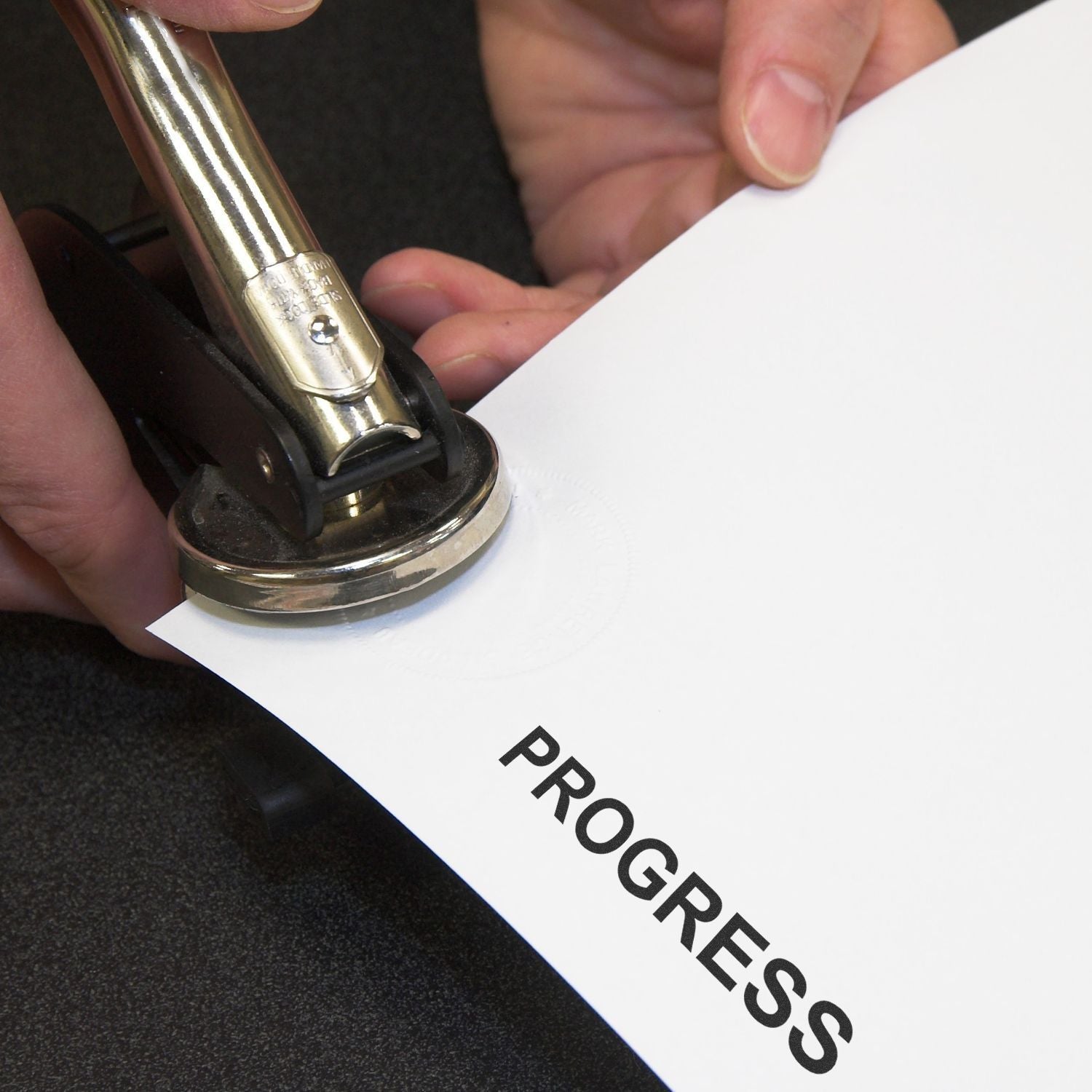 Close-up of a handheld embossing stamp pressing the word PROGRESS into white paper.