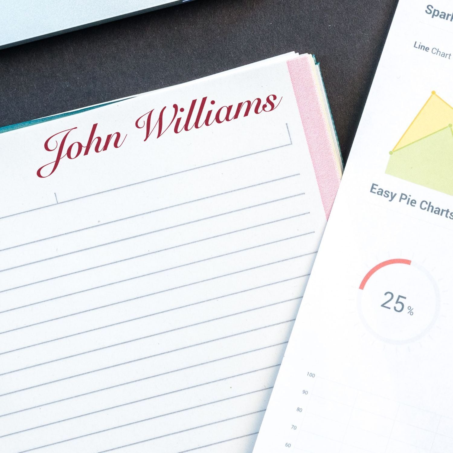 Close-up of personalized notebook with John Williams and business charts featuring line and pie graphs on a desk.