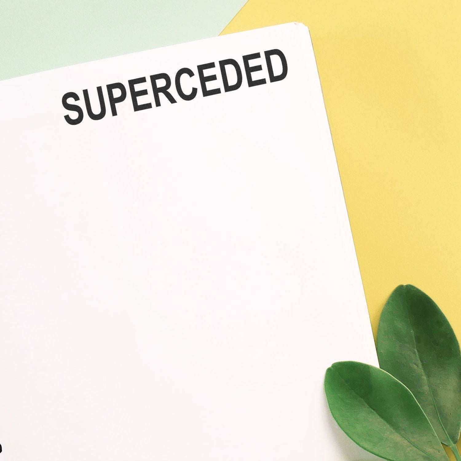 Minimal flat lay of blank white paper with bold SUPERCEDDED text on a pastel yellow background with green leaves.