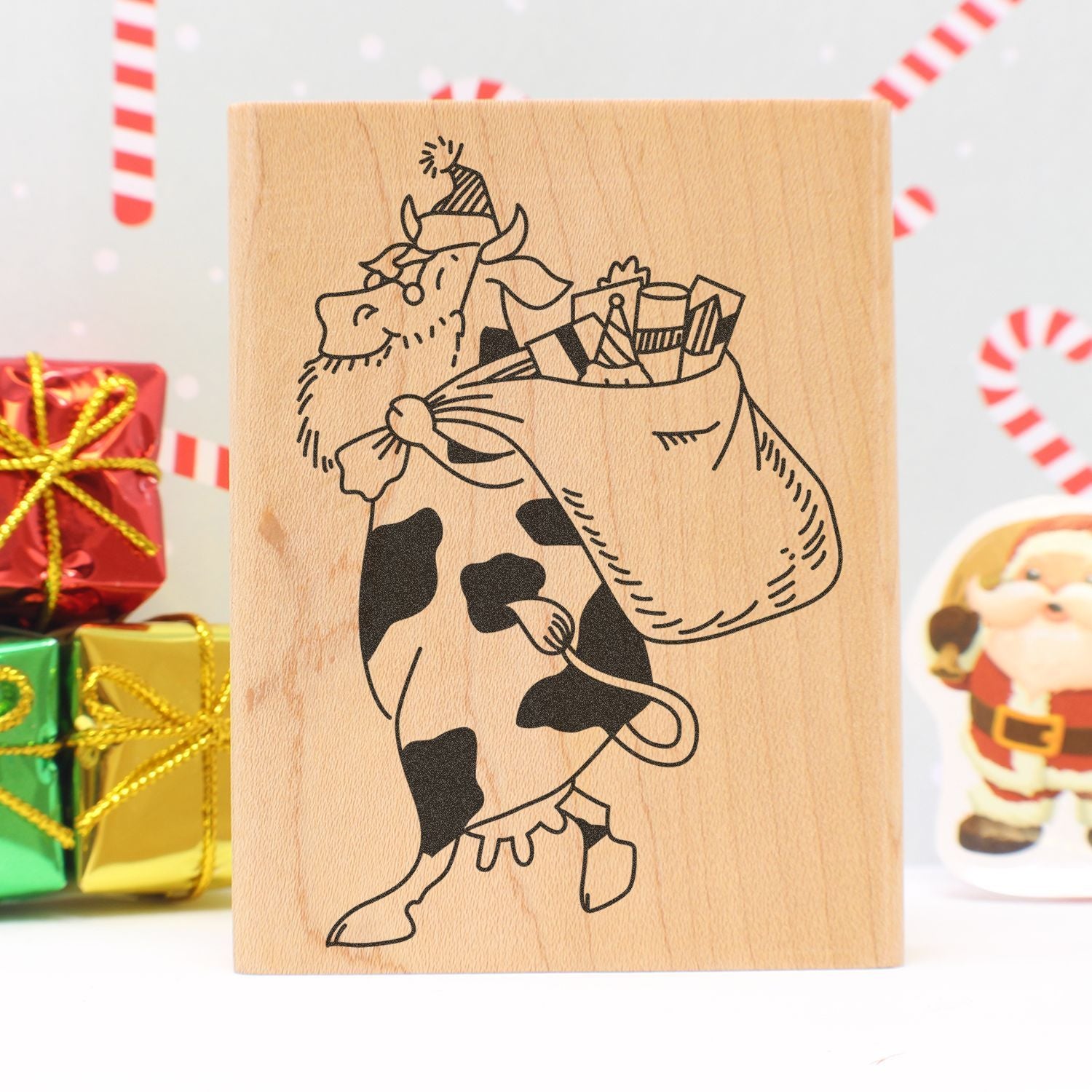 Wooden Christmas stamp featuring a cartoon cow carrying a sack of gifts, with festive holiday presents in the background.