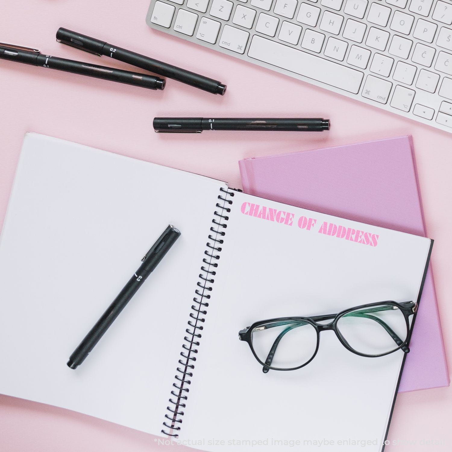Pink stationery flat lay with spiral notebook stamped Change of Address, black pens, eyeglasses, and a white keyboard on a pastel pink desk.