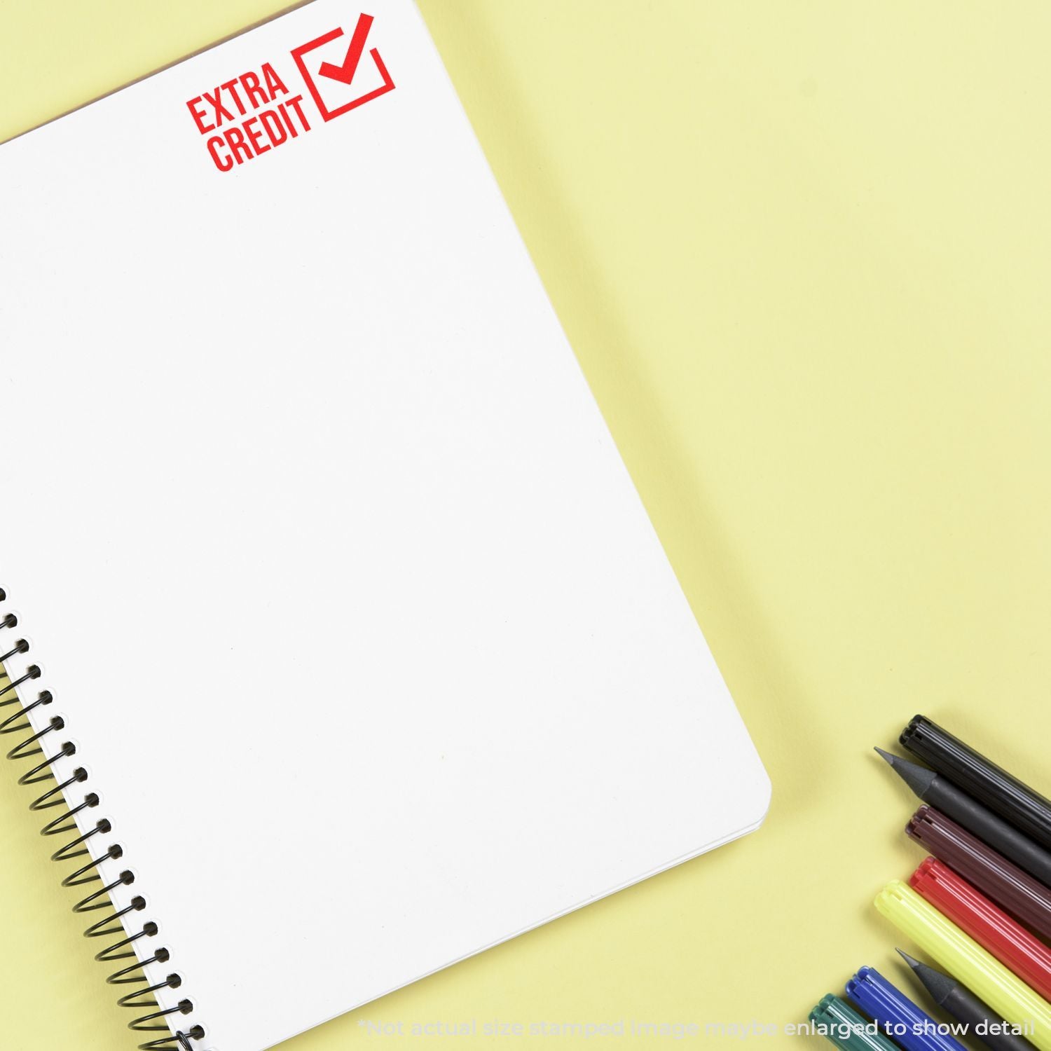 Notebook with Extra Credit stamp and colorful pens on yellow background, promoting study motivation and academic rewards.