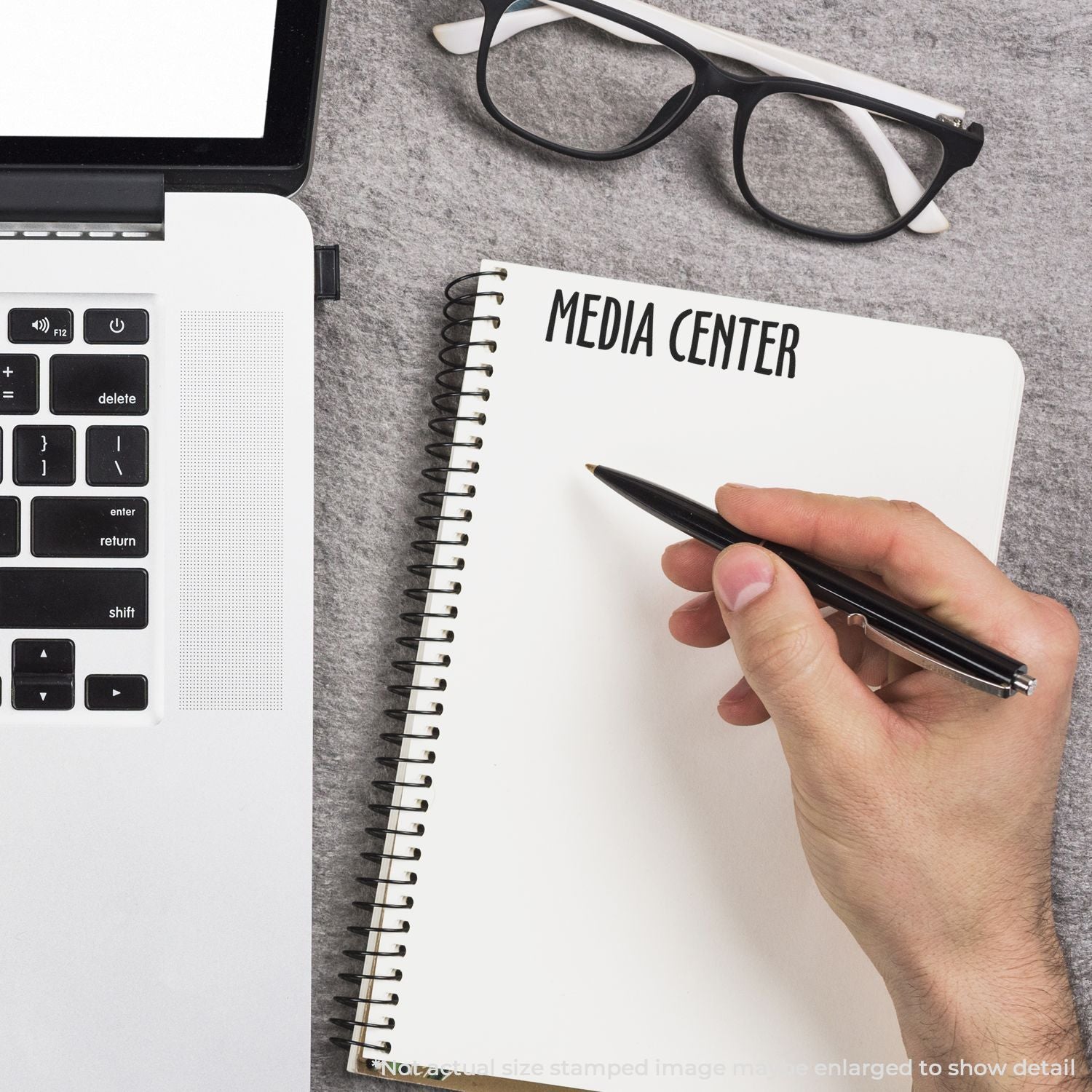Hand writes on notebook 'MEDIA CENTER' beside laptop and eyeglasses, illustrating media management and content creation setup.