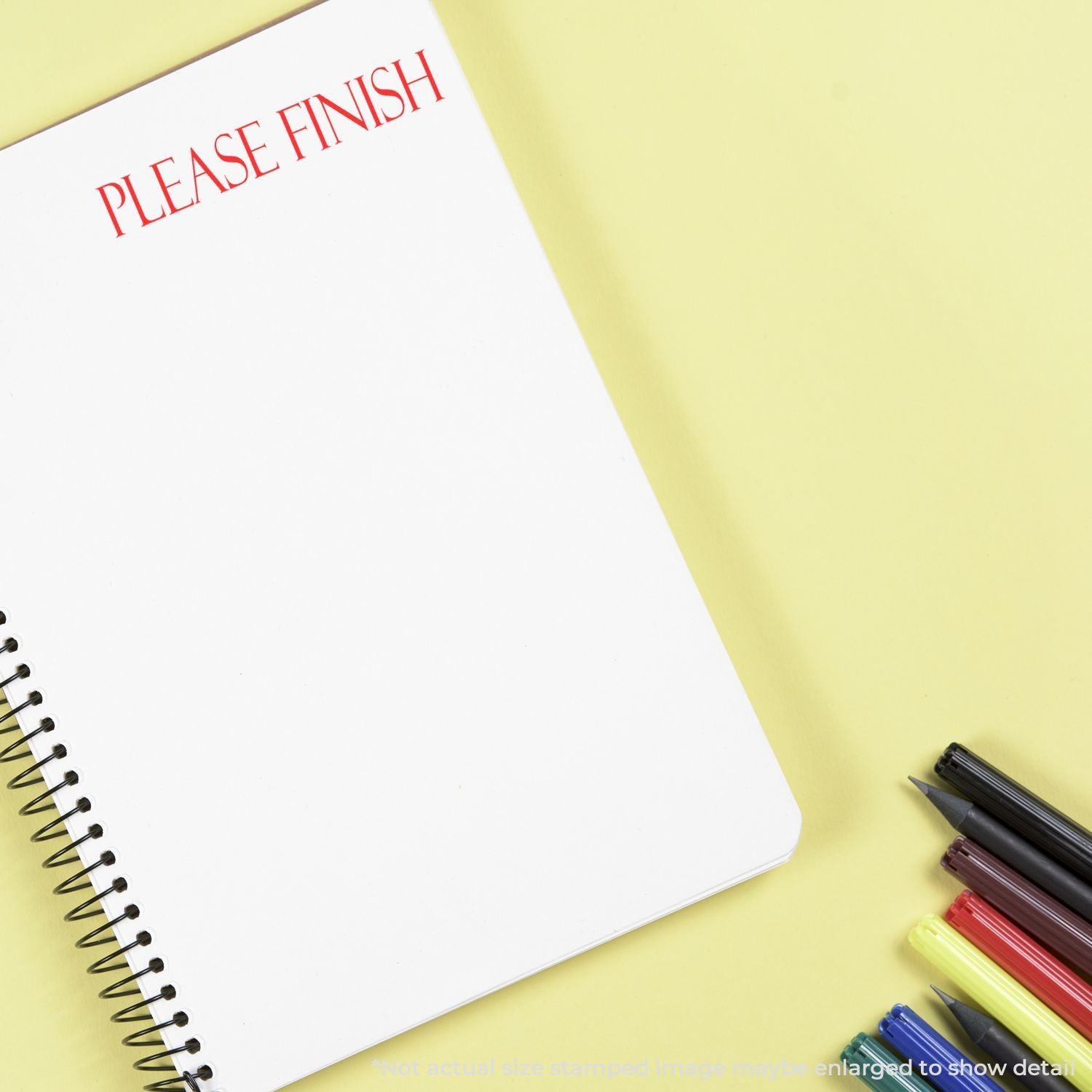 Spiral notebook with PLEASE FINISH text and colorful pens on a yellow background, office stationery concept.