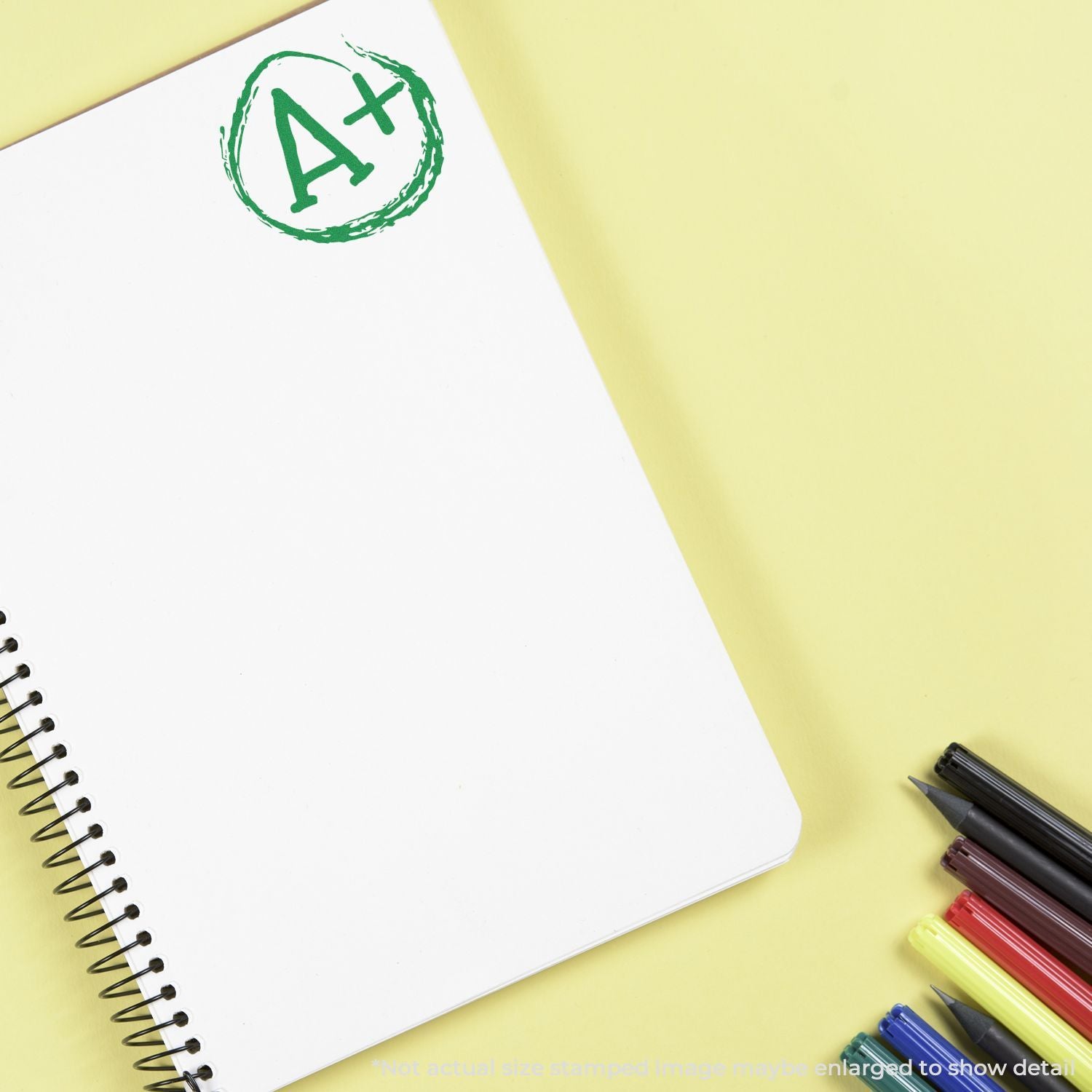 Blank spiral notebook with A+ grade stamp and colorful pens on yellow background, school success and education concept.