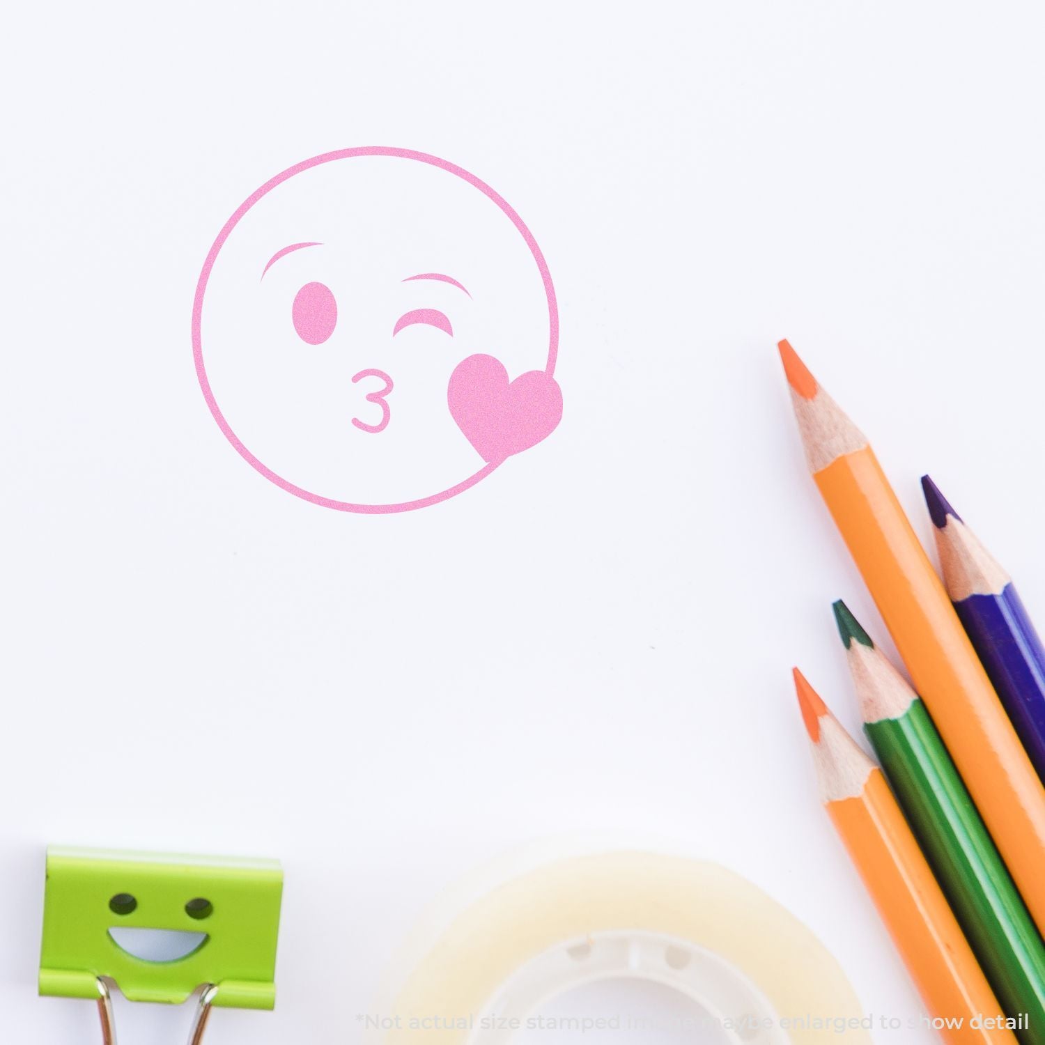 Pink kiss emoji stamp on white paper with colorful pencils and binder clip, creative stationery design.