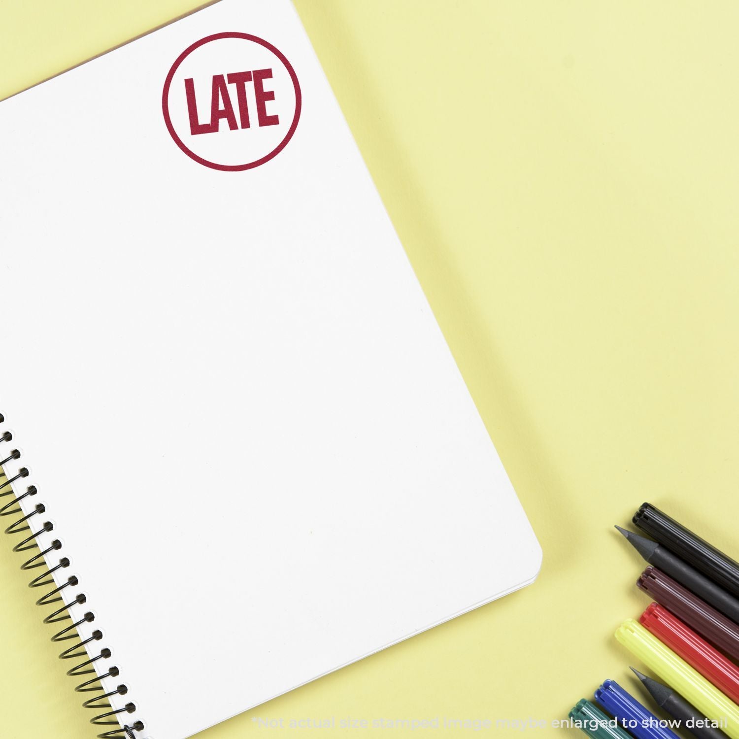 Late stamp on blank spiral notebook page with colorful pens on a yellow background