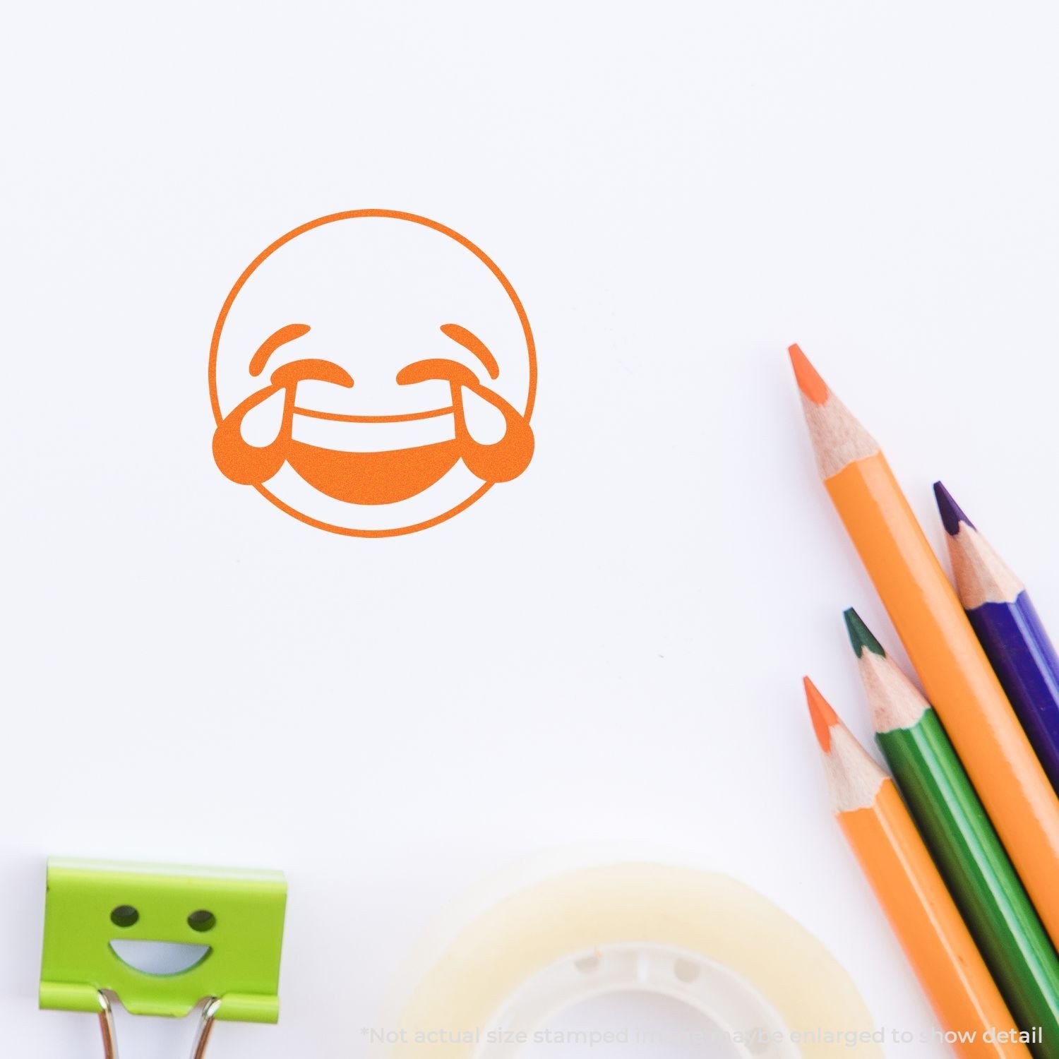 Orange laughing tears emoji stamp on white paper with colorful colored pencils, creative school supplies flat lay.