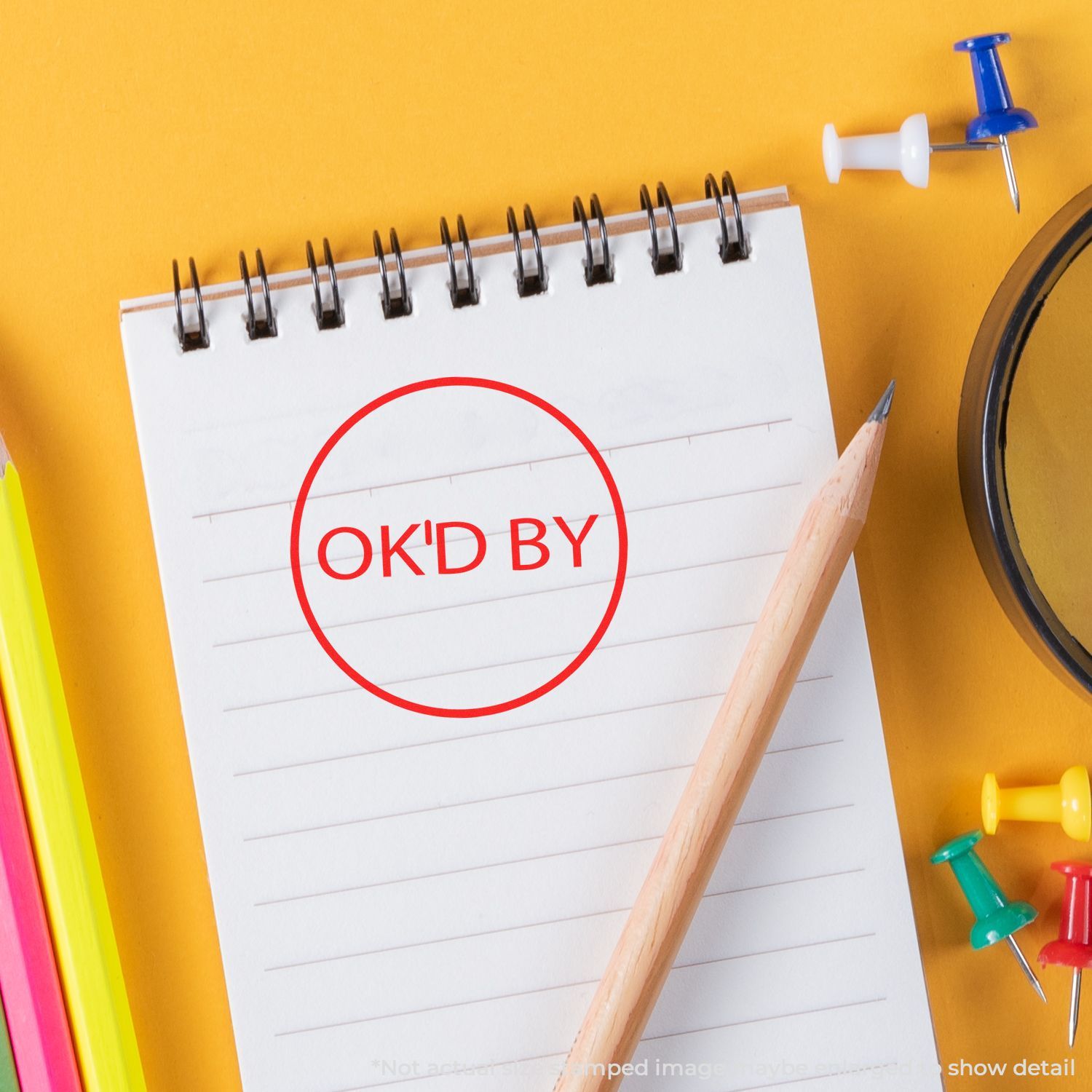 Spiral notebook with red OKD BY approval stamp, pencil, and colorful stationery on a yellow background.