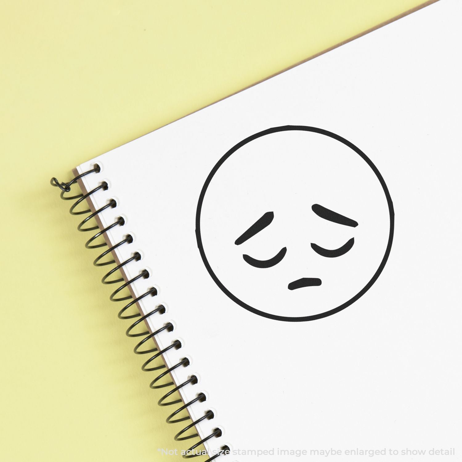 Spiral notebook with sad face icon on white page against a pale yellow background, symbolizing sadness or stress.