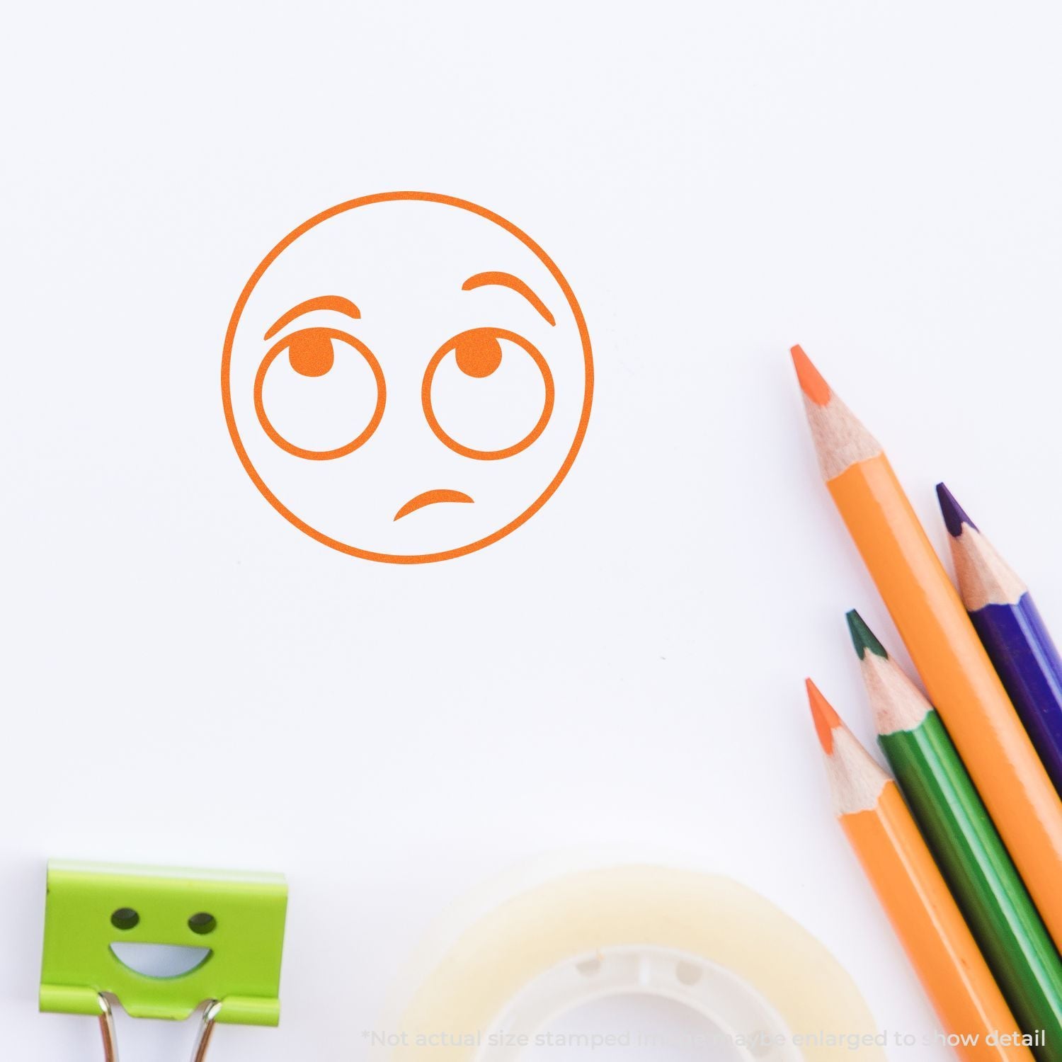 Orange sad face emoji stamp on white paper with colored pencils and a smiling binder clip, creative kids art supplies.