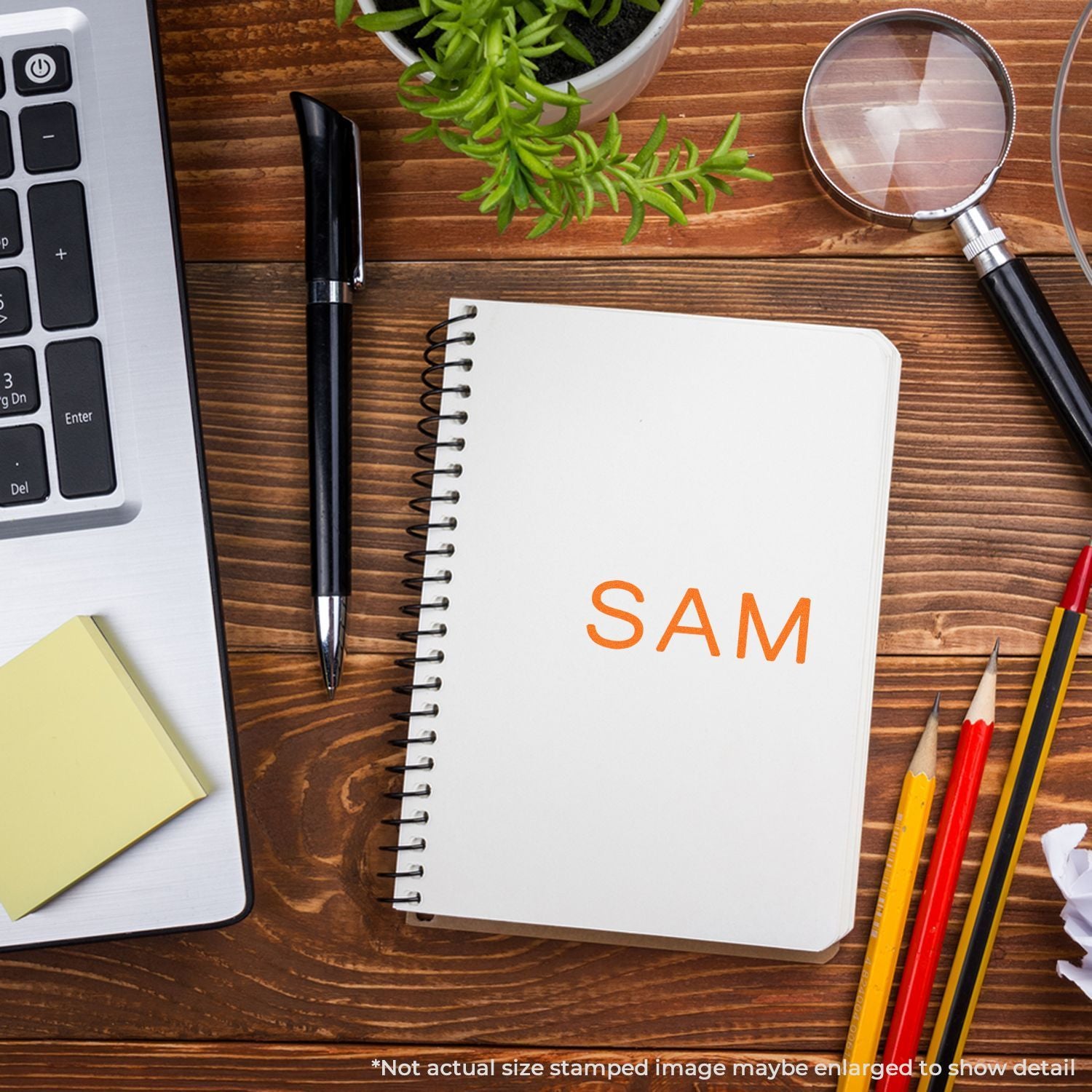 Top view of a wooden office desk flat lay with a spiral notebook labeled SAM, laptop, pen, magnifying glass, pencils, and a potted plant.