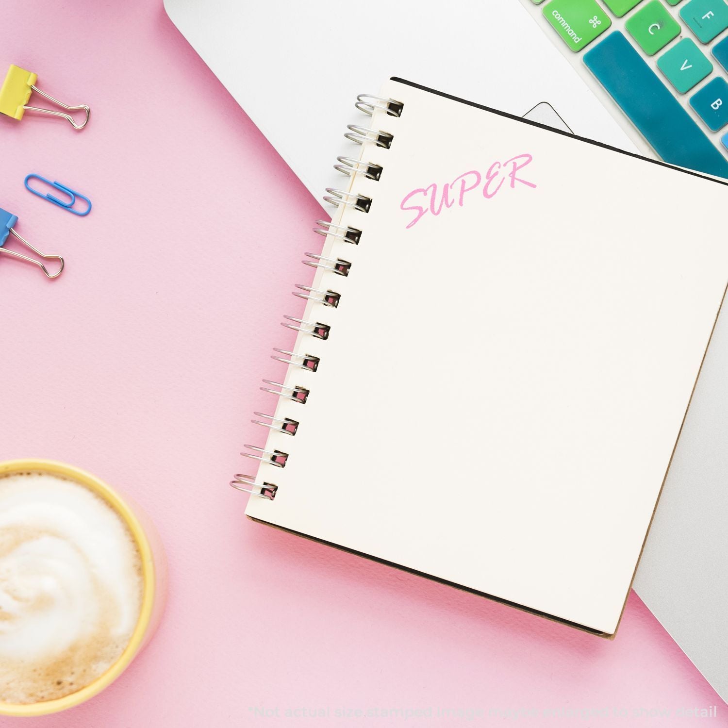Pastel flat lay of spiral notebook with SUPER text, coffee cup, colorful keyboard, and paper clips on a pink workspace.