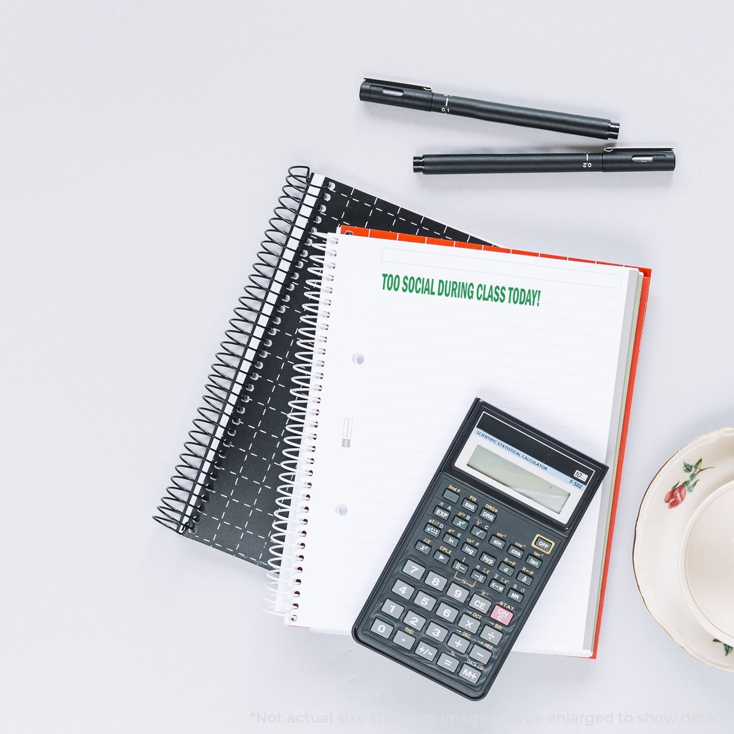 Top view of school stationery with notebooks, scientific calculator, pens, and coffee cup on a white desk, featuring a notebook that says Too social during class today!
