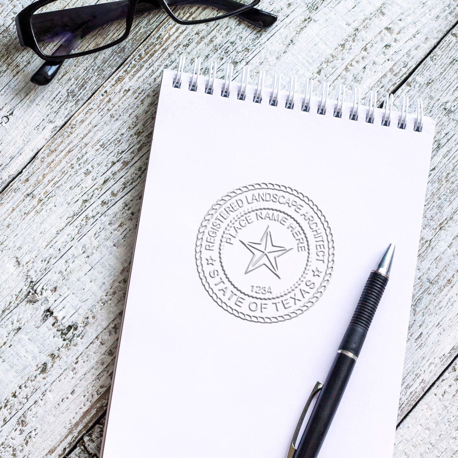 Notepad with Texas registered landscape architect seal, pen, and eyeglasses on rustic wood desk.