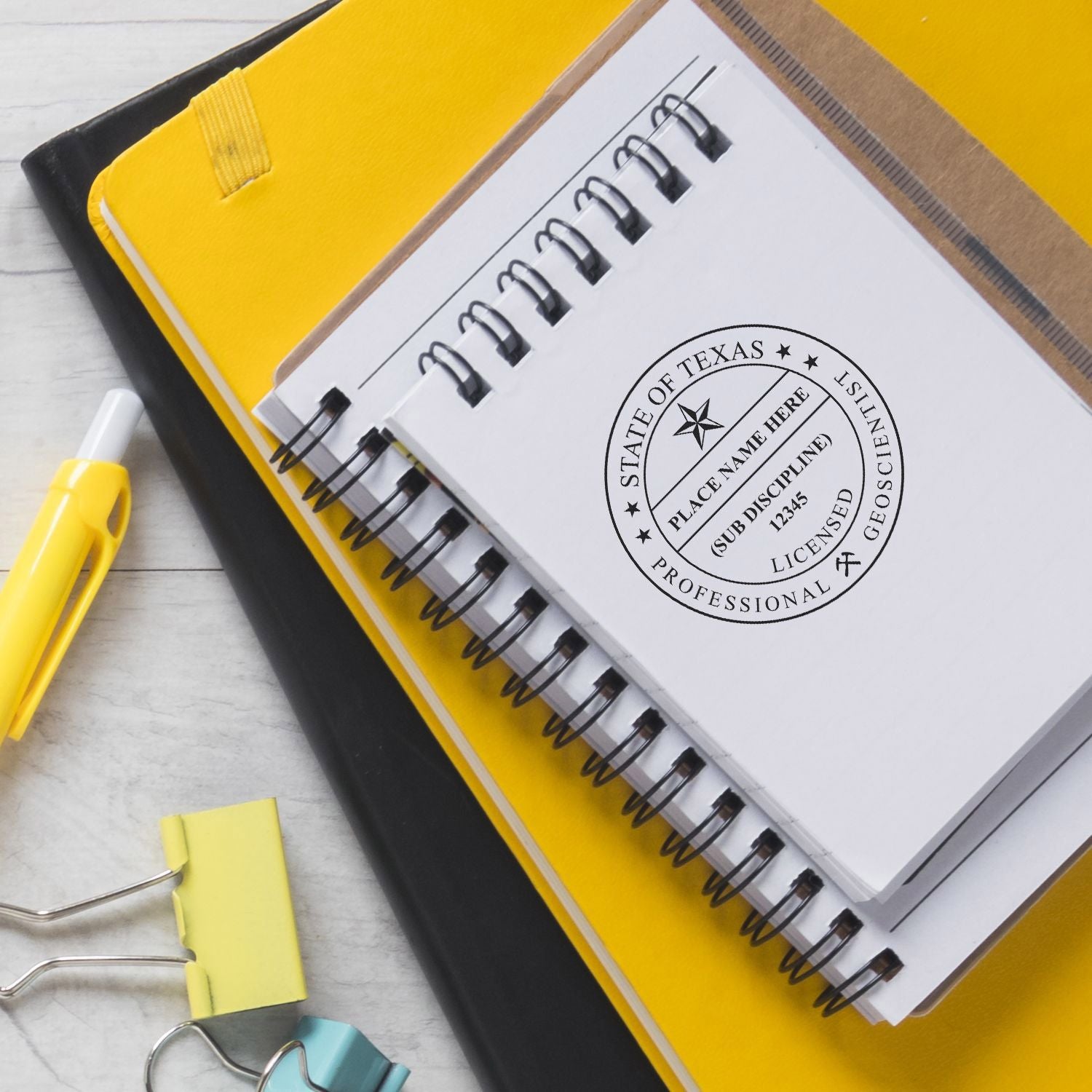 Yellow spiral notebook with State of Texas professional geoscientist seal, office stationery, and binder clips on a desk.