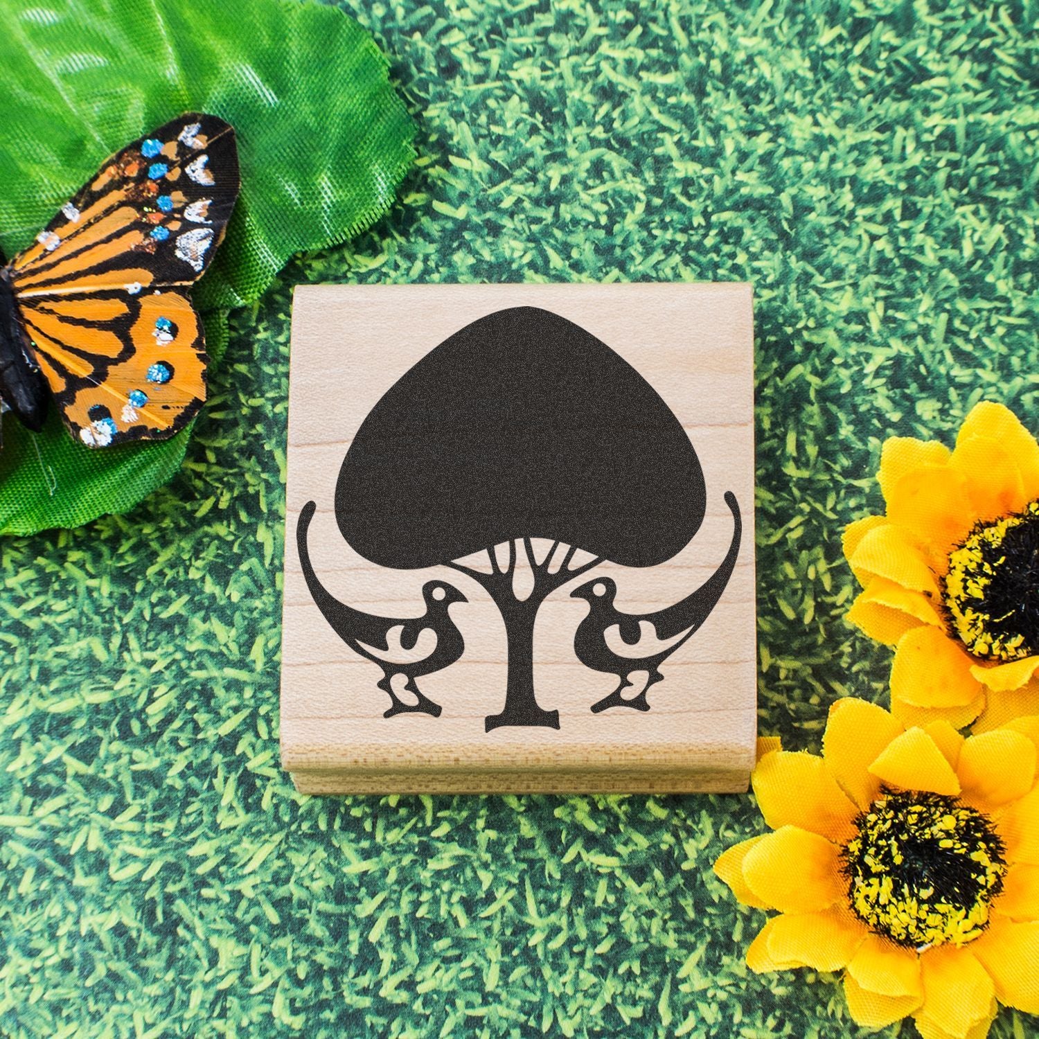 Decorative wooden rubber stamp with black tree-and-birds logo, butterfly, and sunflower on a green nature background.