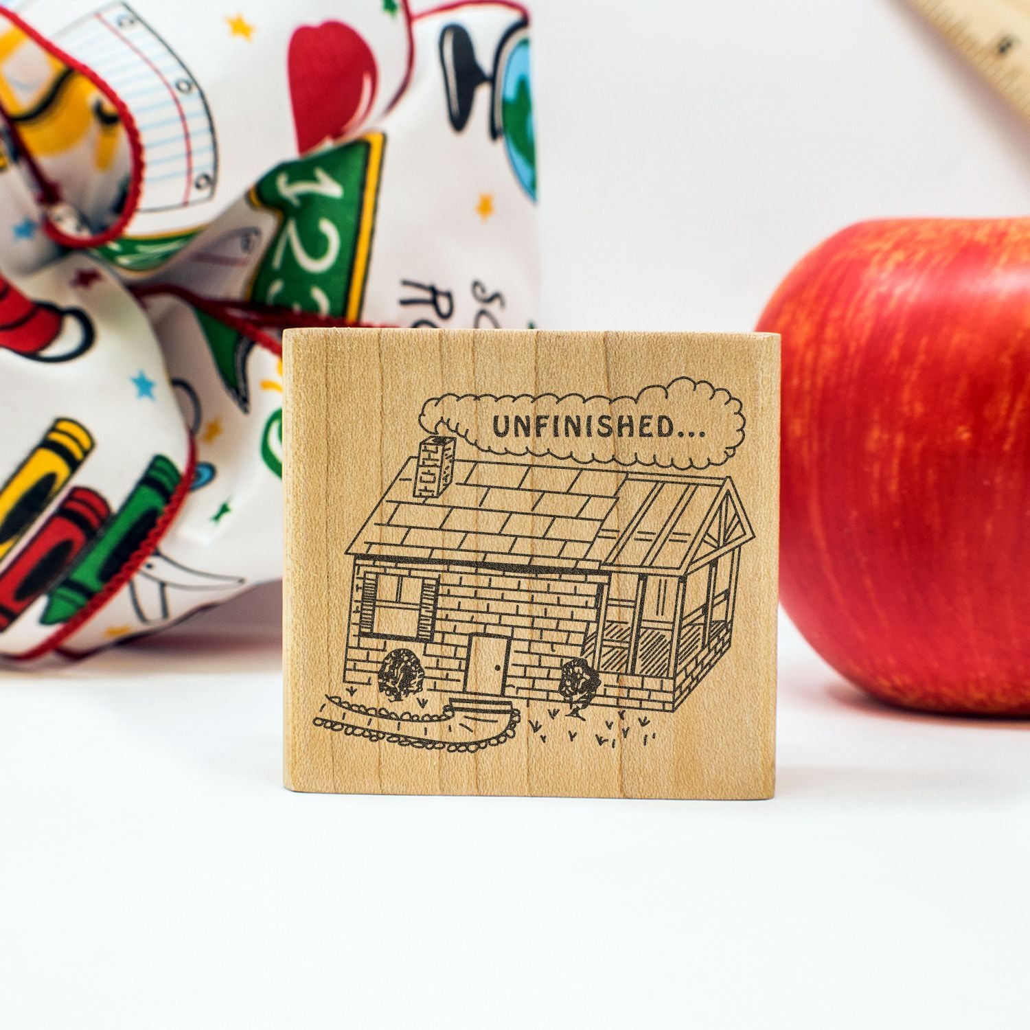 Wooden rubber stamp with unfinished house illustration, red apple, and colorful school-themed background.