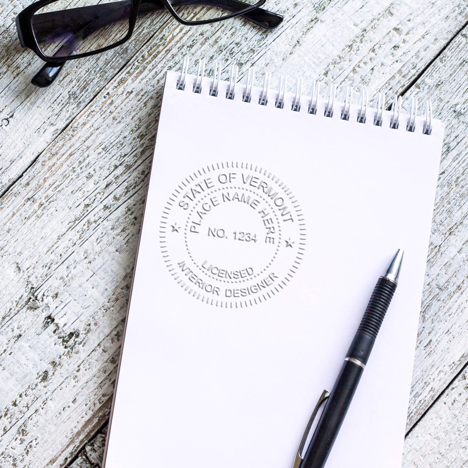 Blank spiral notebook with embossed Vermont licensed interior designer logo, pen, and eyeglasses on rustic wood desk.