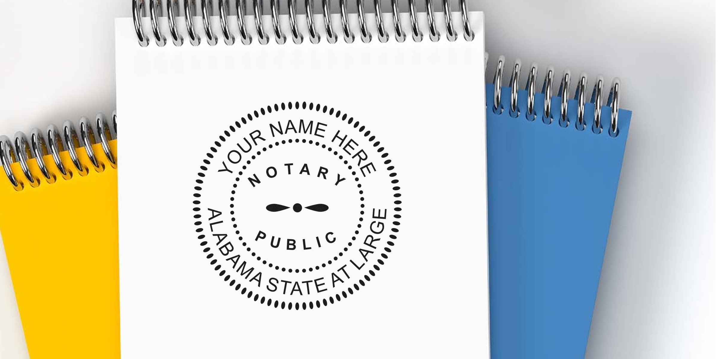 Alabama notary seal expiration and compliance shown on a custom stamp on a white spiral notebook, with yellow and blue notebooks in the background.