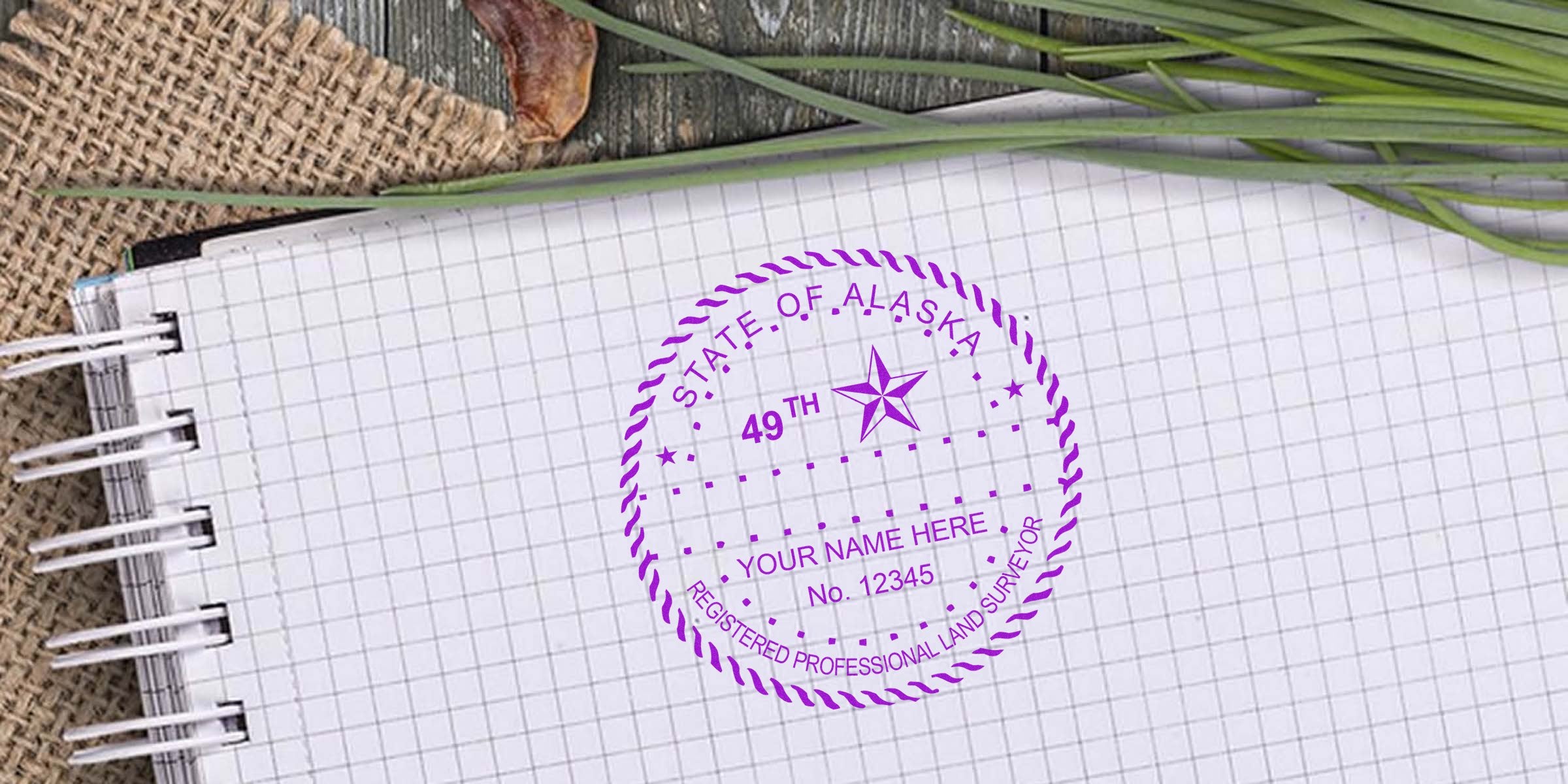 Alaska land surveyor stamp on graph paper notebook with green grass and burlap background, featuring a professional stamp available for purchase.