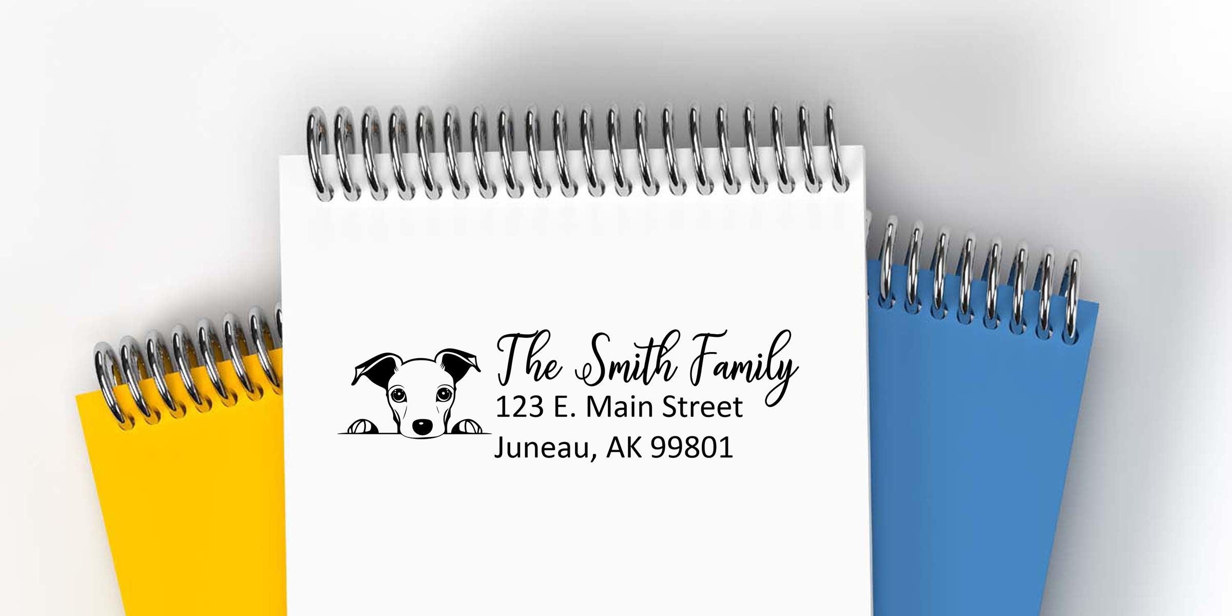 Custom spiral notebook with dog logo and personalized family address reading “The Smith Family, 123 E. Main Street, Juneau, AK 99801,” shown with yellow and blue notebooks in the background