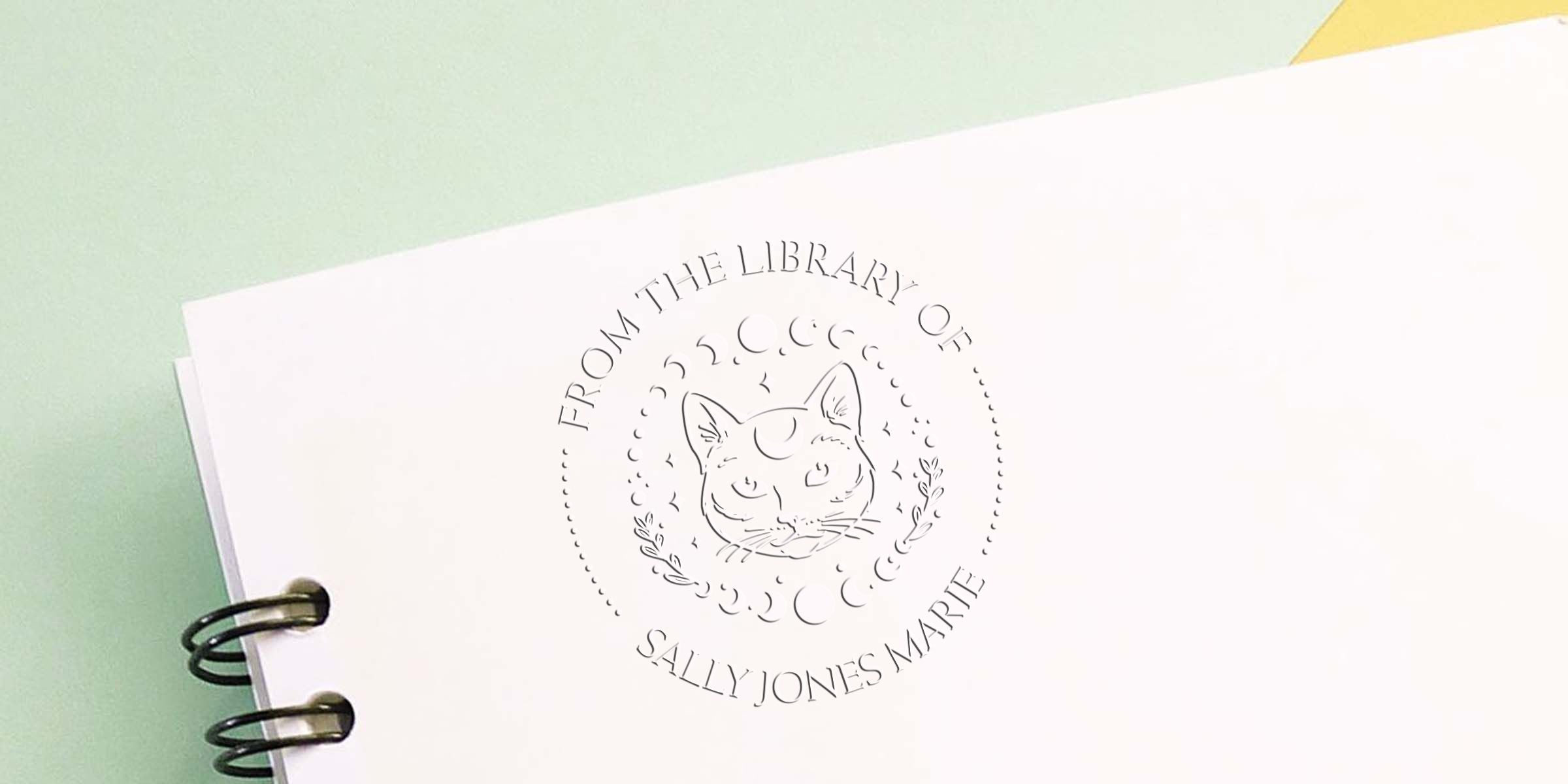 Minimalist branded notebook cover with spiral binding, featuring a cat logo and the text “From the Library of Sally Jones Marie” on a pastel green and white background