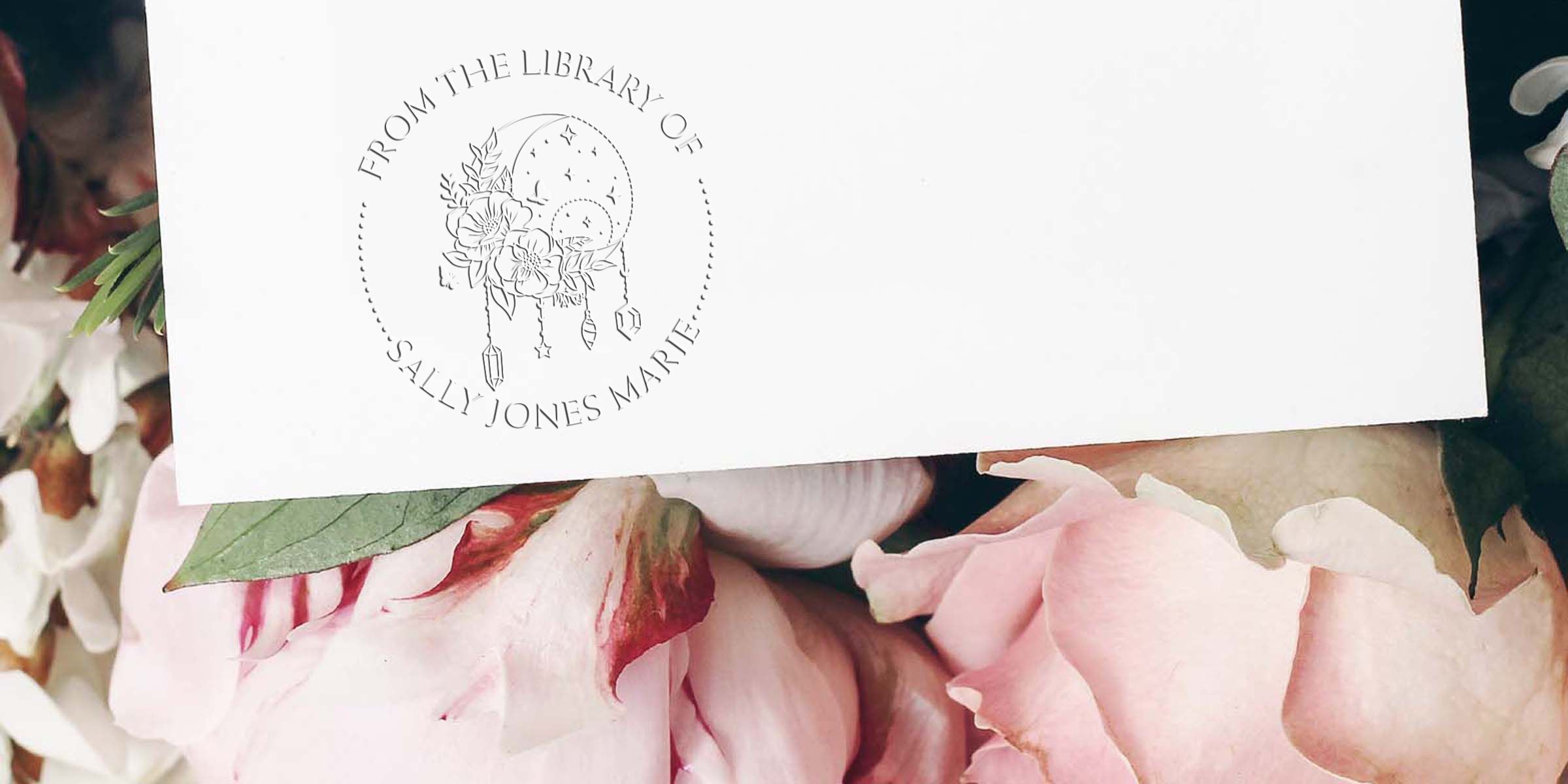 White personalized stationery card with a floral dreamcatcher logo reading “From the Library of Sally Jones Marie” resting on pastel pink flowers and greenery
