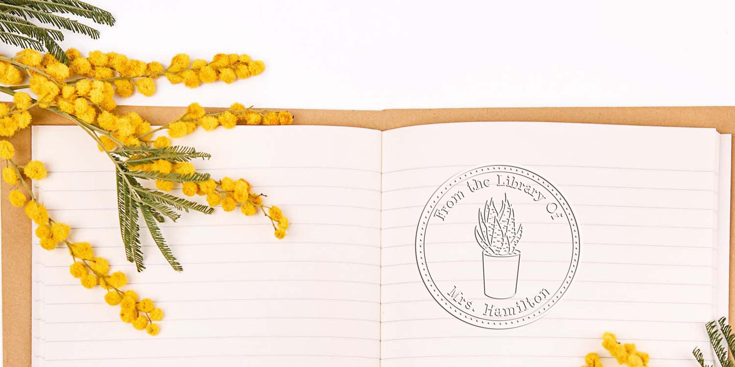 Open lined notebook with yellow mimosa flowers and green fern leaves arranged around a botanical library logo reading “From the Library of Mrs. Hamilton”