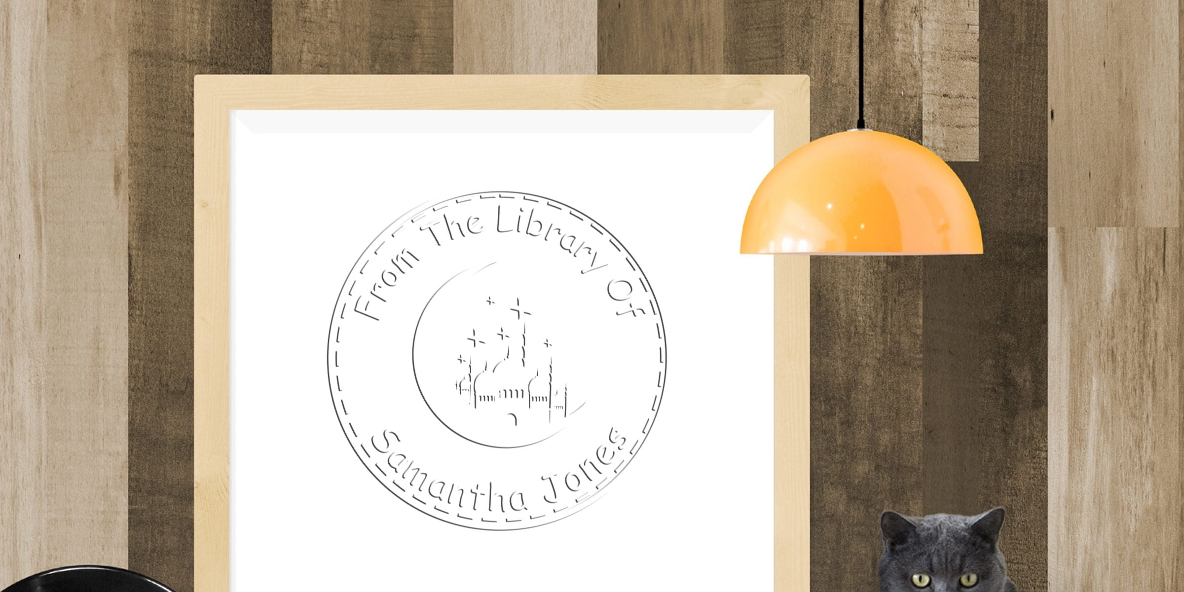 Framed personalized library stamp wall art reading “From the Library Of Samantha Jones” in a modern wood-paneled room with an orange pendant lamp and gray cat