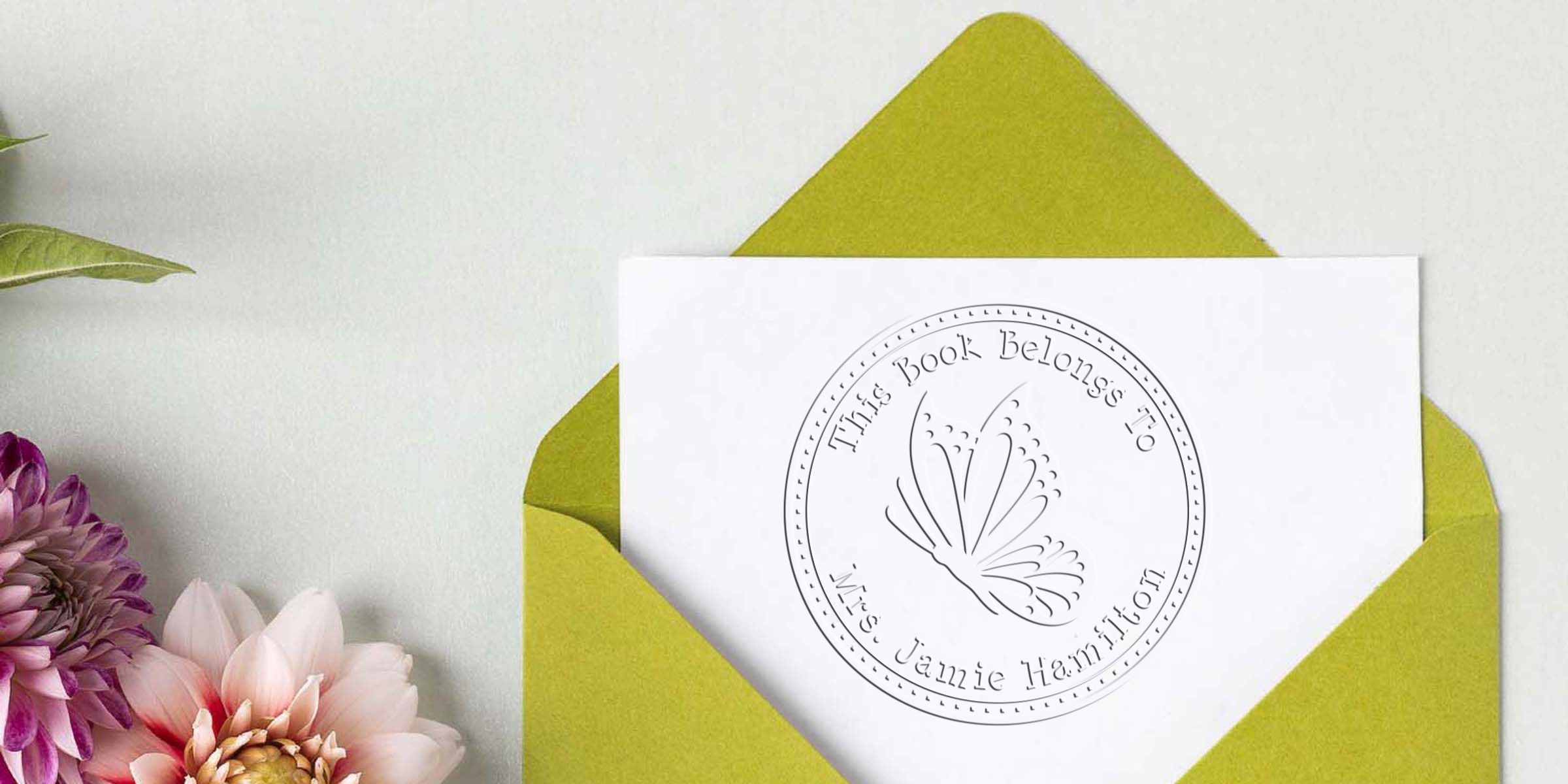 White personalized bookplate card in a lime green envelope, featuring a butterfly logo and the text “This Book Belongs To Mrs. Jamie Hamilton,” styled with floral accents on a light background