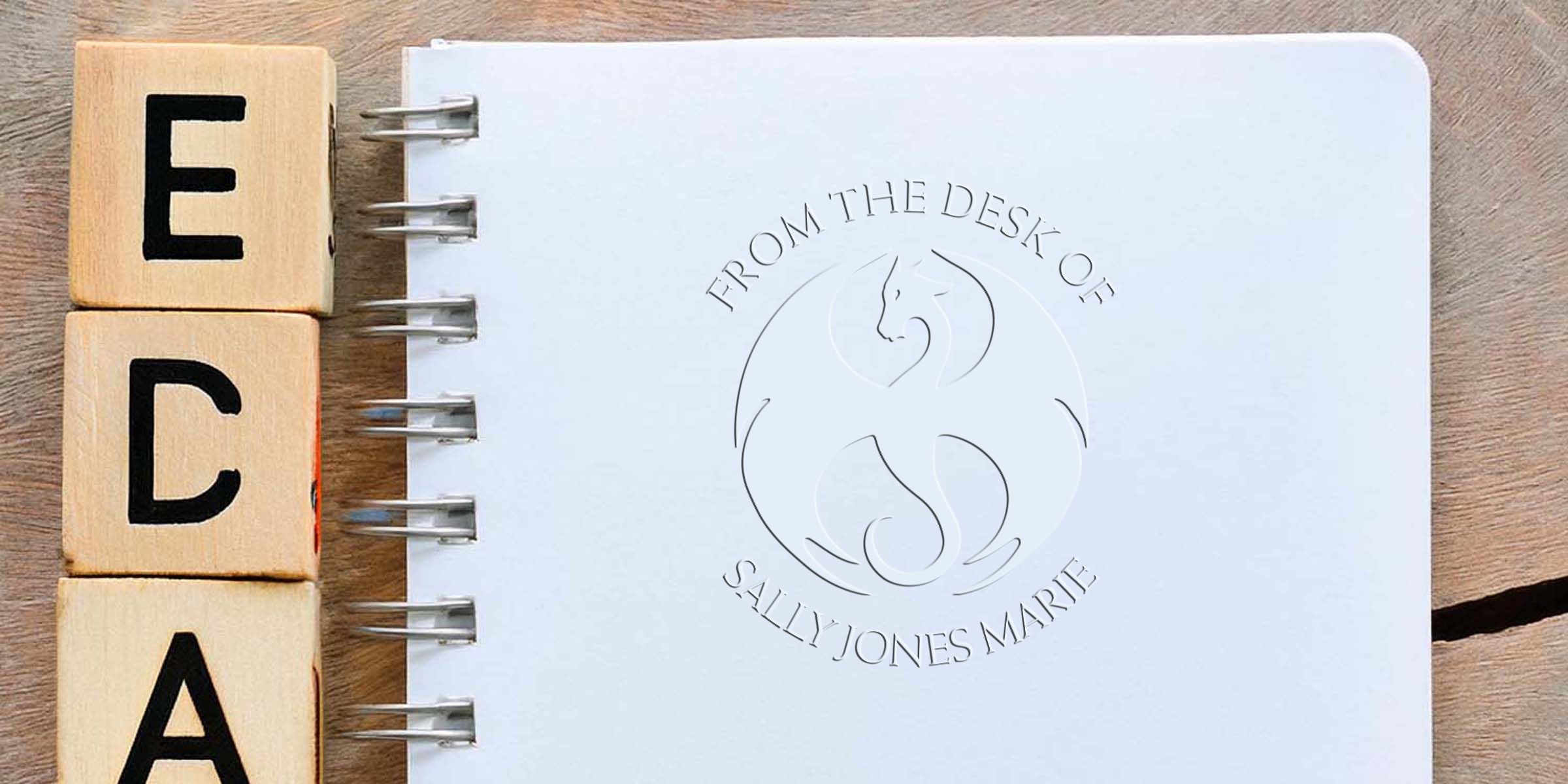 Wooden alphabet blocks spelling EDA beside a spiral notebook with an embossed “From the Desk of Sally Jones Marie” logo on a wooden tabletop, custom stationery branding design