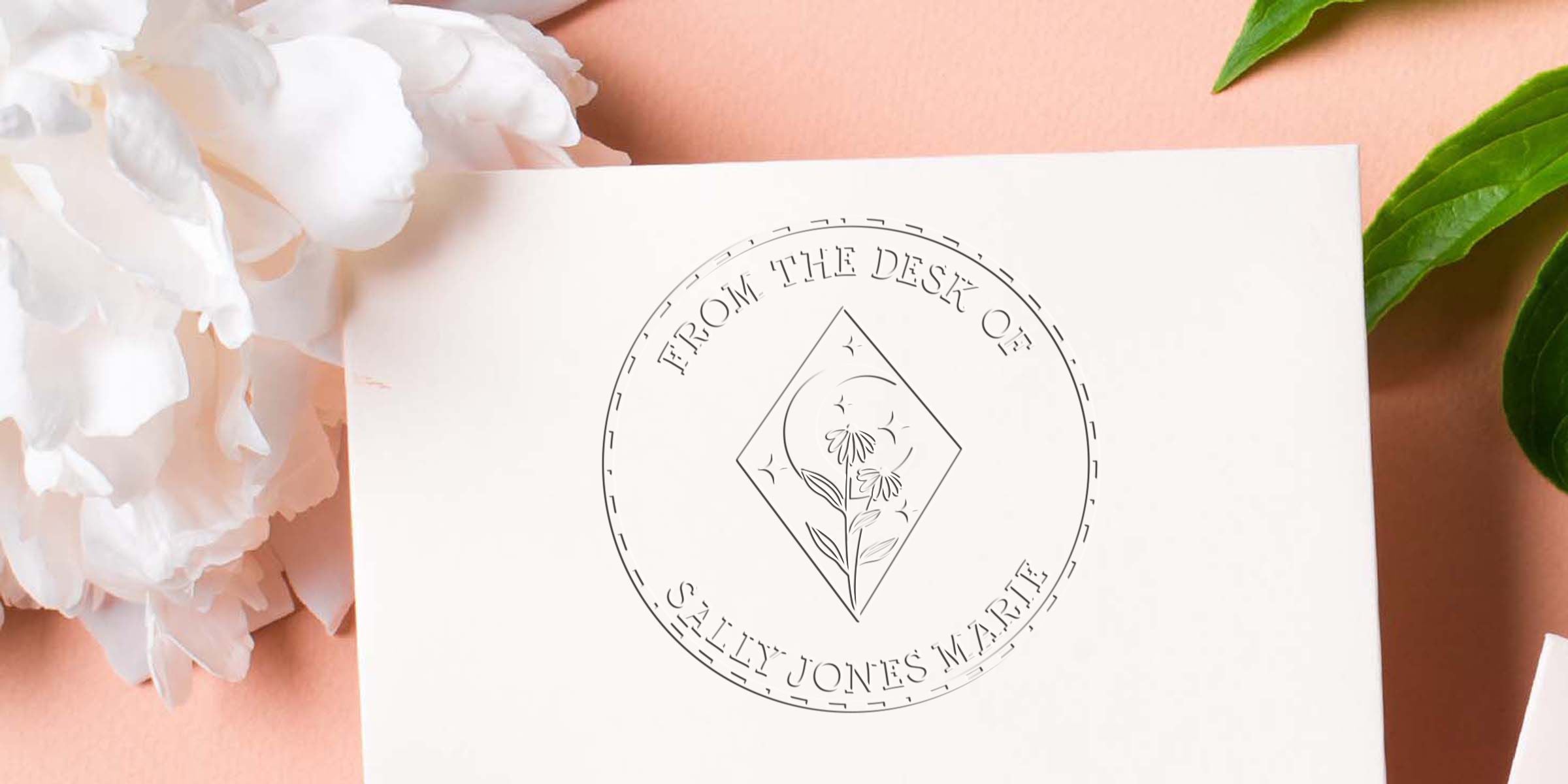 Elegant custom stationery flat lay with a minimalist floral logo reading “From the Desk of Sally Jones,” on a soft peach background with white flowers and green leaves