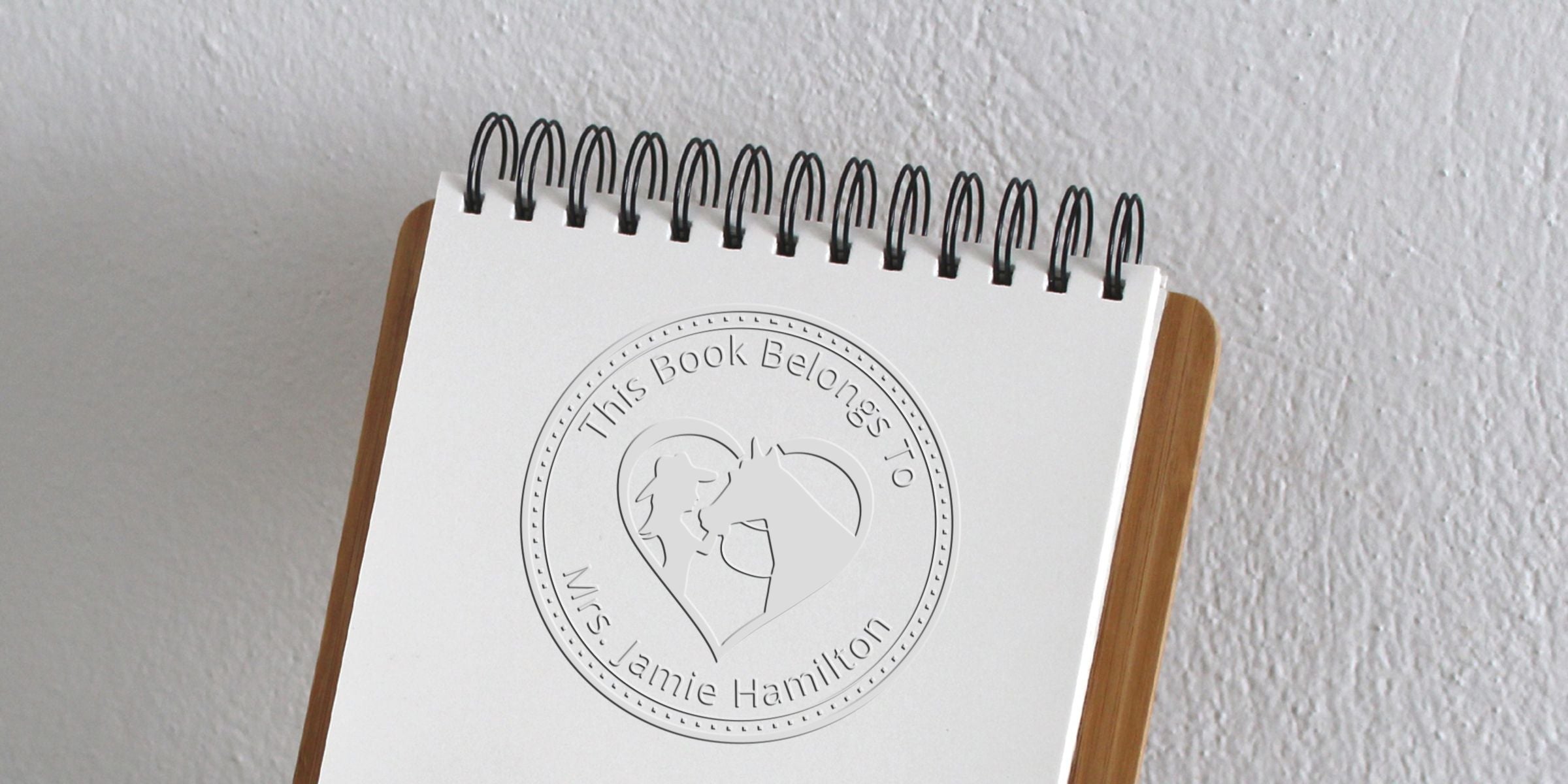 Personalized spiral-bound notebook on a wooden stand with embossed “This Book Belongs To Mrs. Jamie Hamilton” cover and heart-shaped design against a white wall