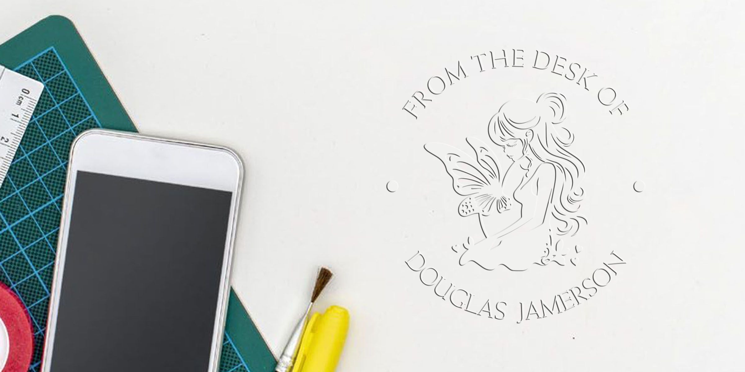 Custom engraved “From the Desk of Douglas Jamerson” logo with fairy illustration on a crafting workspace, beside a smartphone, cutting mat, ruler, and paintbrush