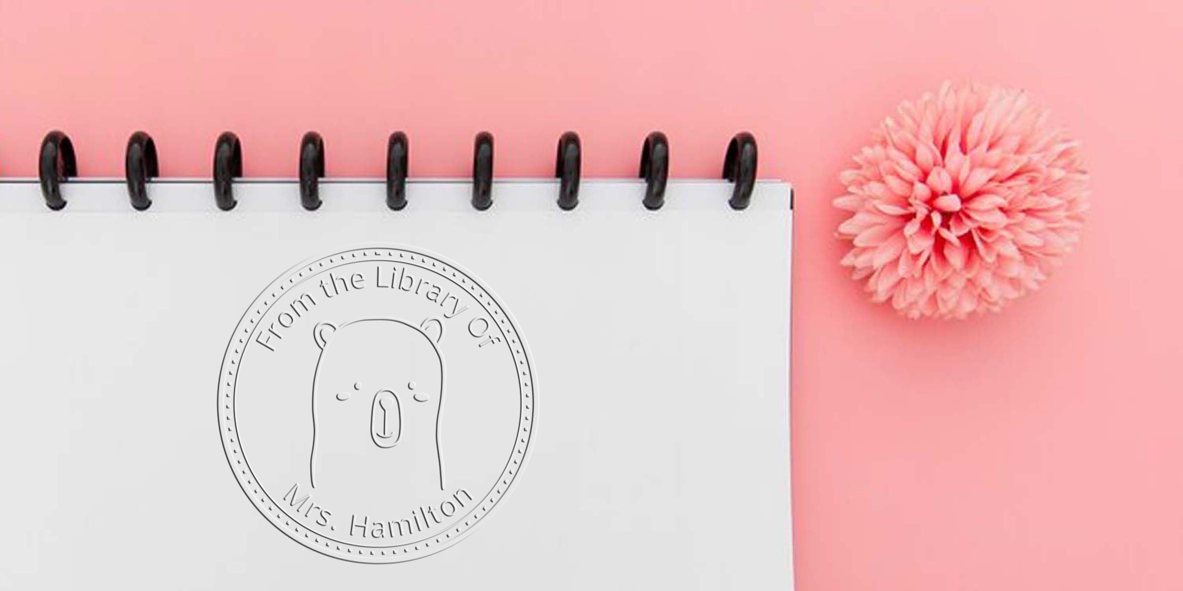 Minimalist white spiral notebook with “From the Library of Mrs. Hamilton” bear embossing, arranged on a pink background with a decorative pink flower, personalized stationery and book stamp design