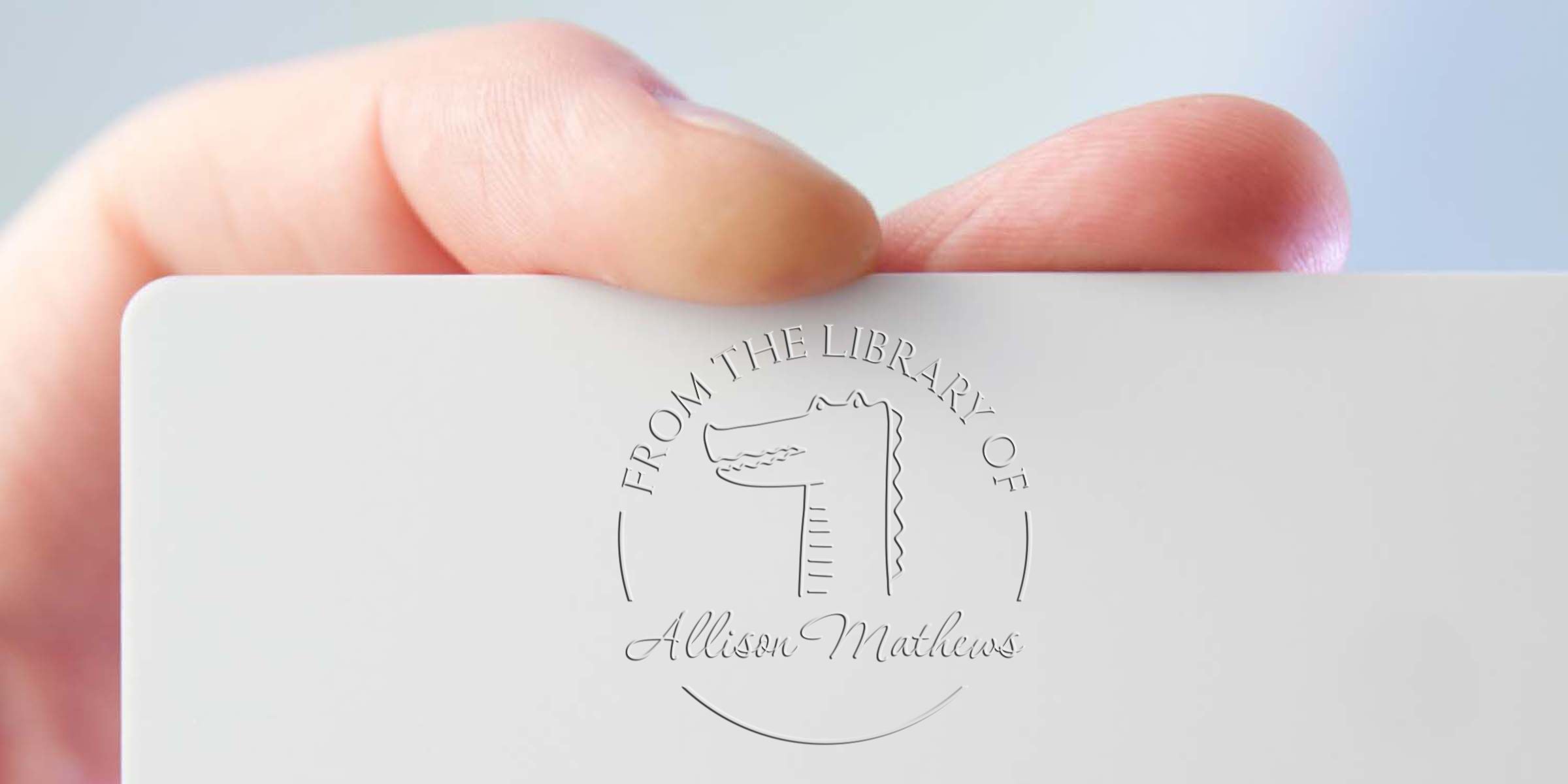 Close-up of a hand holding a white card with embossed “From the Library of Allison Mathews” bookplate logo against a soft blue background
