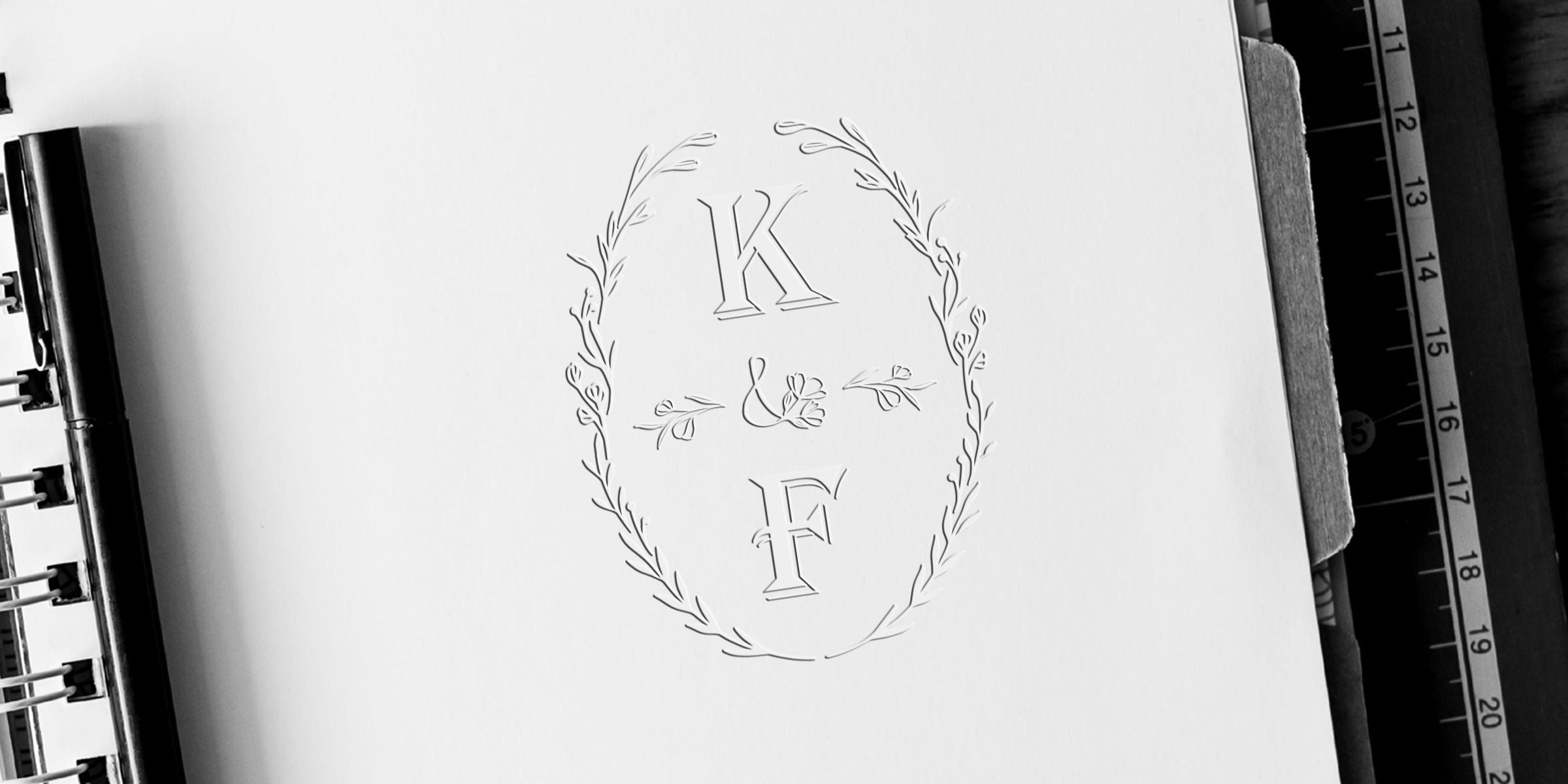 Elegant black and white K and F monogram sketch inside a delicate floral wreath on notebook paper, with spiral binding and ruler visible рядом