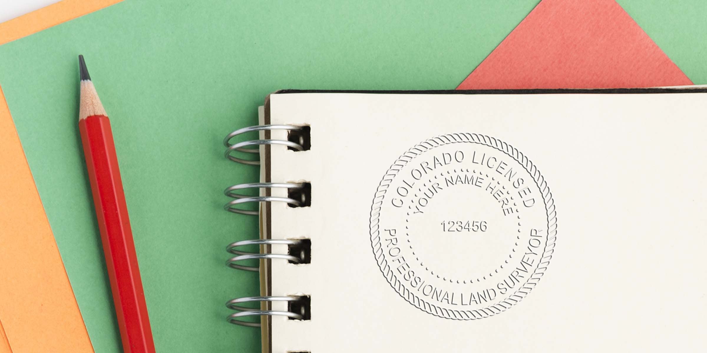 Colorado land surveyor stamp on notebook with red pencil, displaying stamp and size requirements on a colorful paper background.