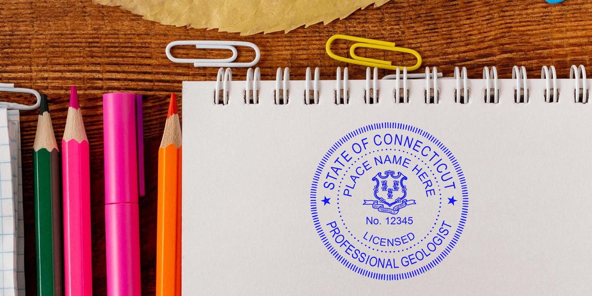 Licensed professional geologist stamp on white spiral notebook with colorful pencils and paper clips on wooden desk, highlighting Connecticut geologist stamp cost and fees.