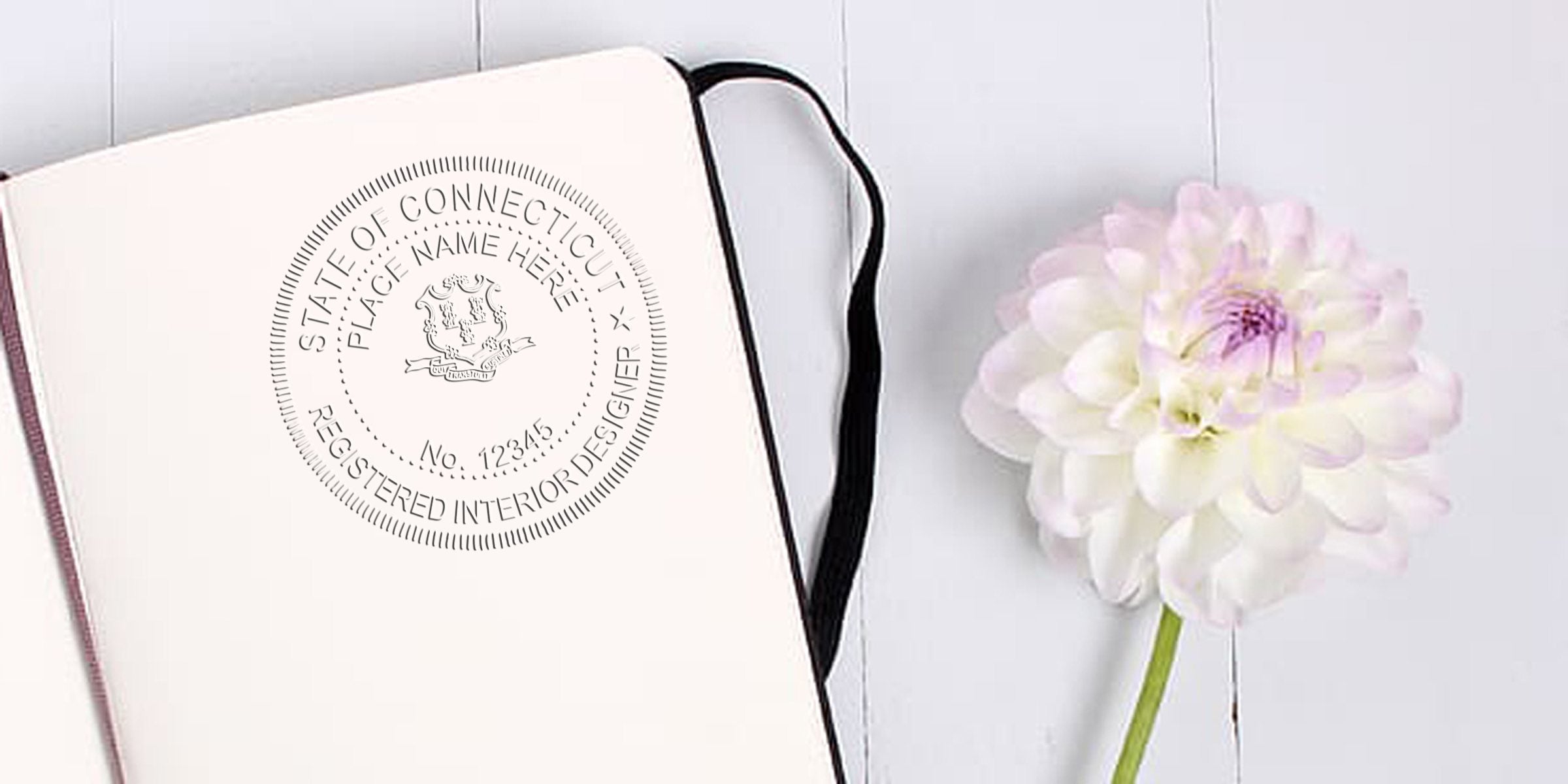 Open notebook showing Connecticut registered interior designer seal with specifications, beside a white flower on a white wooden surface.