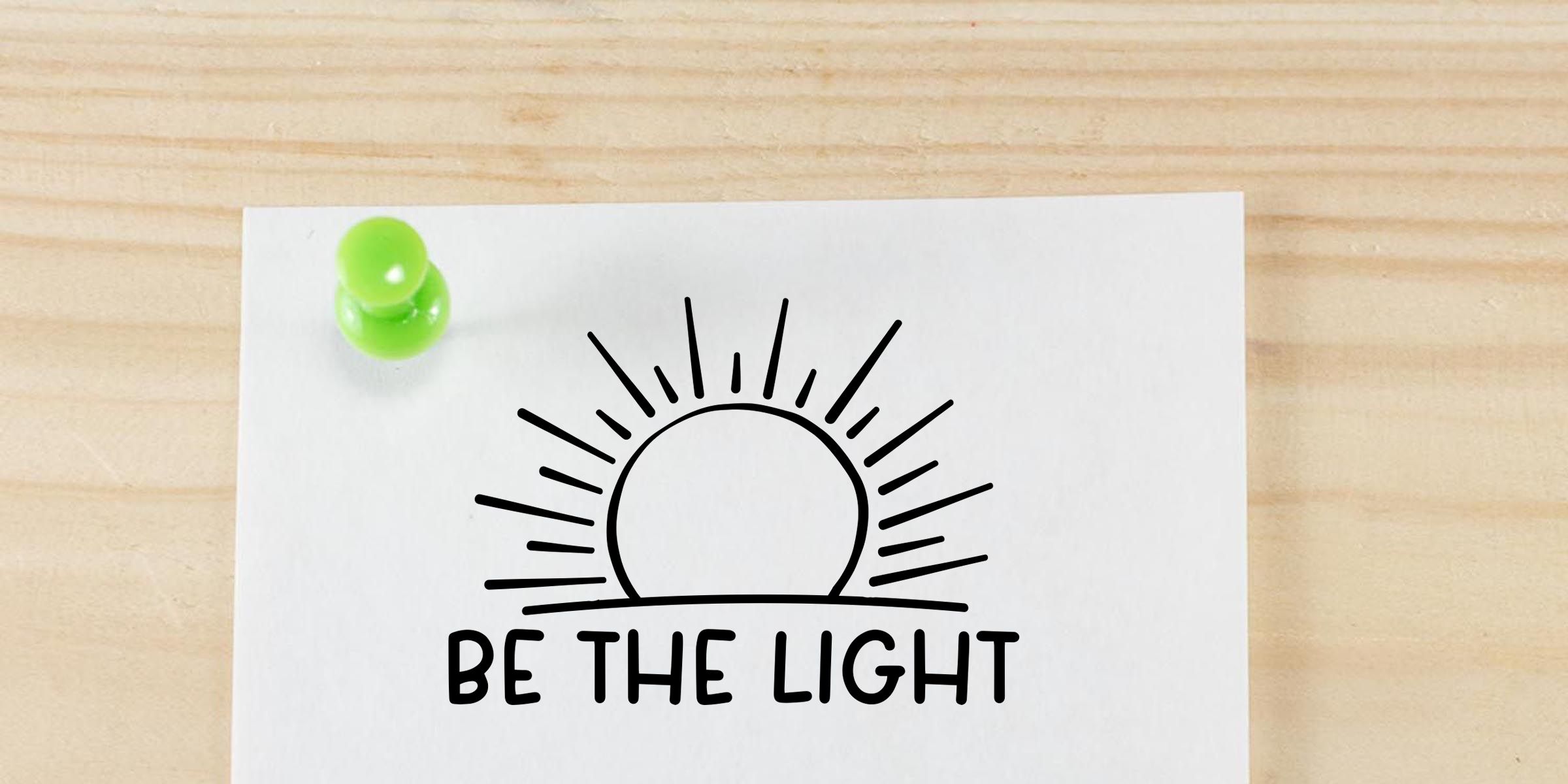 White motivational card pinned on a wooden board with a green pushpin, featuring a sunburst icon and the phrase “Be the Light”