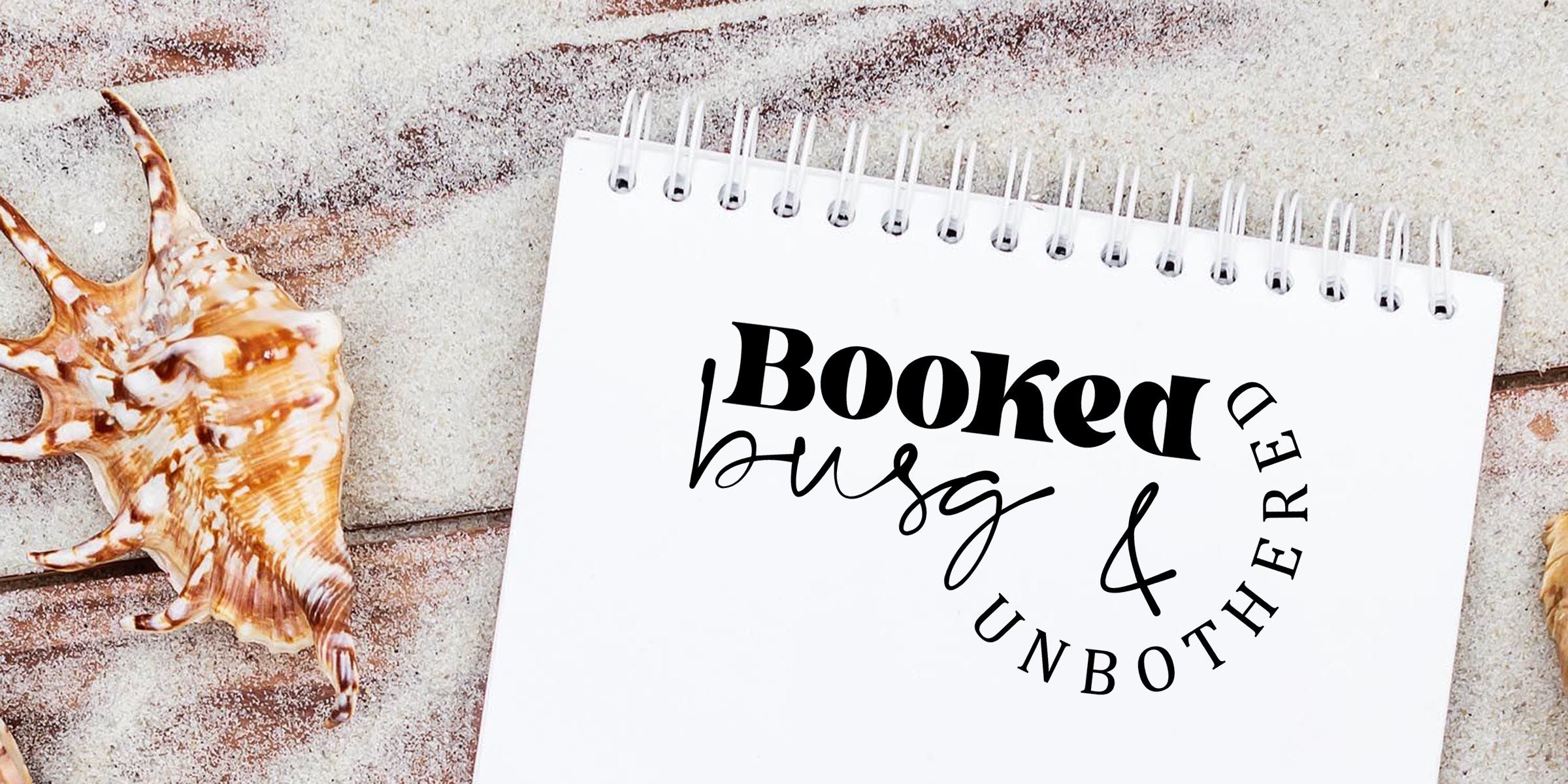 Spiral notebook with “Booked busy & unbothered” typography on a sandy beach background with seashells.