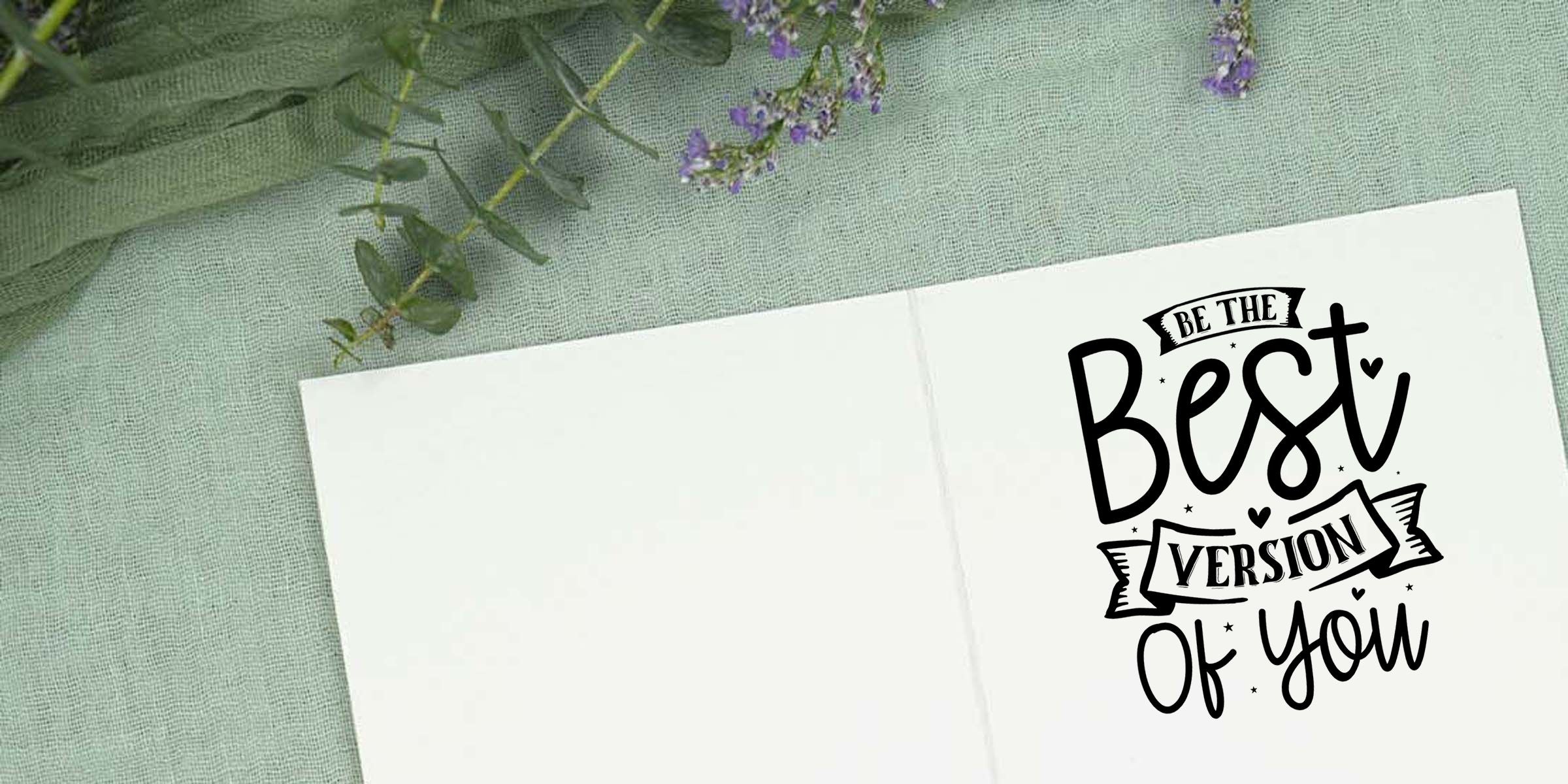 Open notebook on green textured fabric with eucalyptus leaves and purple flowers, featuring hand-lettered quote “Be the Best Version of You” on the right page